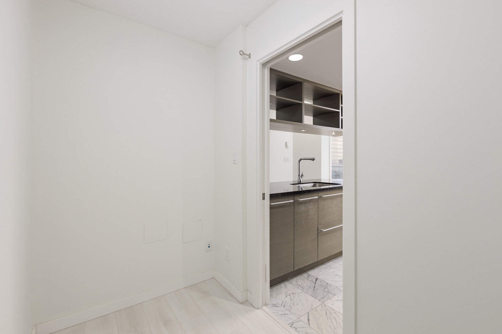 A small, empty white room with light wood flooring leads to a modern kitchen with grey cabinets, a sink, and marble floor tiles.