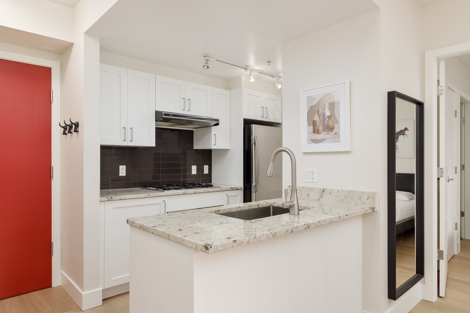 Modern kitchen with white cabinets, granite countertop, stainless steel appliances, and a red door; a bedroom is visible through an open door.