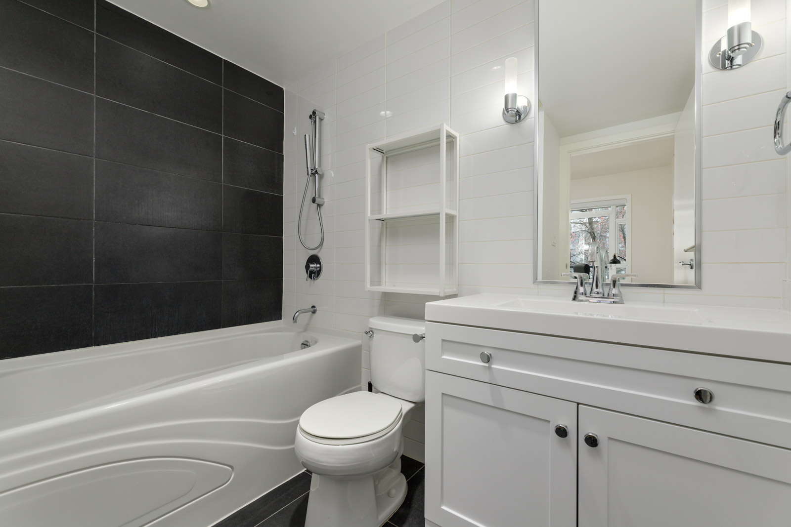 Modern bathroom with a white bathtub, toilet, vanity, shelving, and white tile walls; features a dark accent wall behind the bathtub and chrome fixtures.