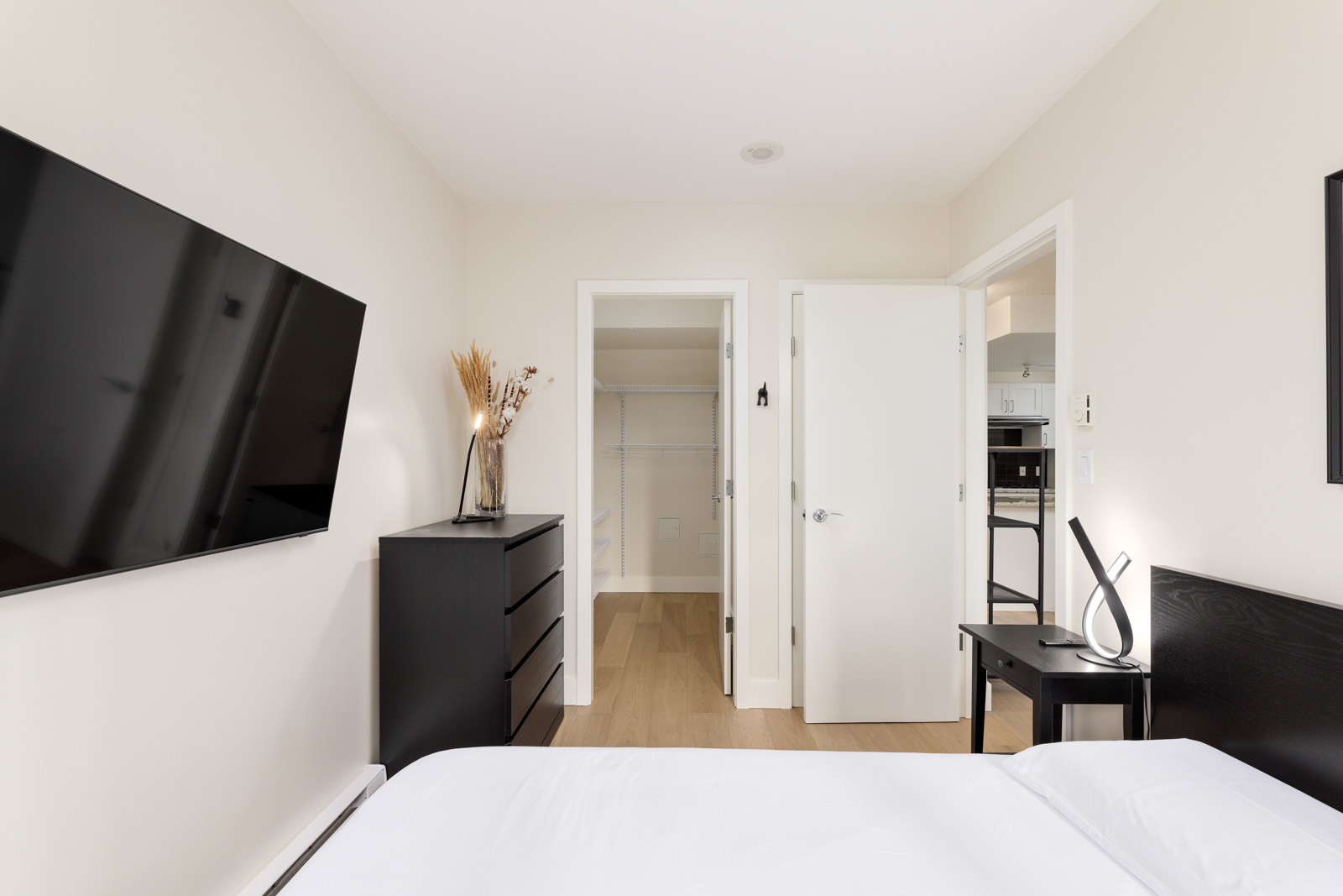 Minimalist bedroom with a wall-mounted TV, black dresser with a vase, open closet door, adjacent open door, and part of a bed and nightstand visible.