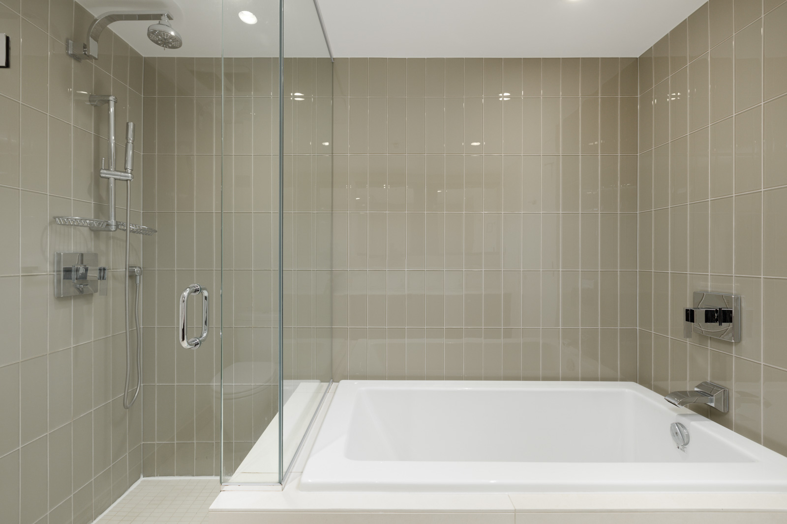 Modern bathroom with beige tiled walls, a glass-enclosed shower on the left, and a white rectangular bathtub on the right.