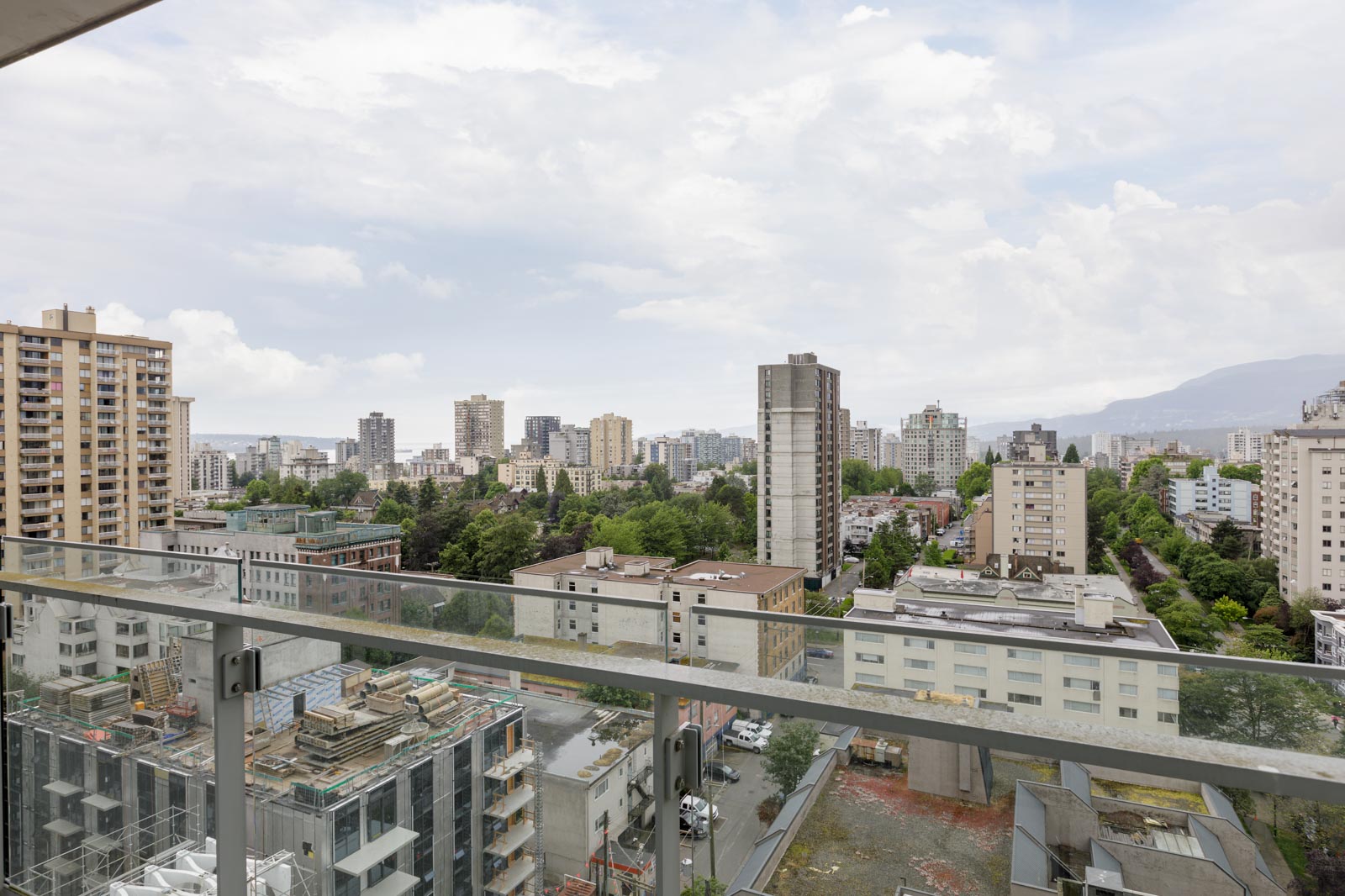 High-rise cityscape view from a balcony, showing apartment buildings, trees, and construction sites under a cloudy sky.
