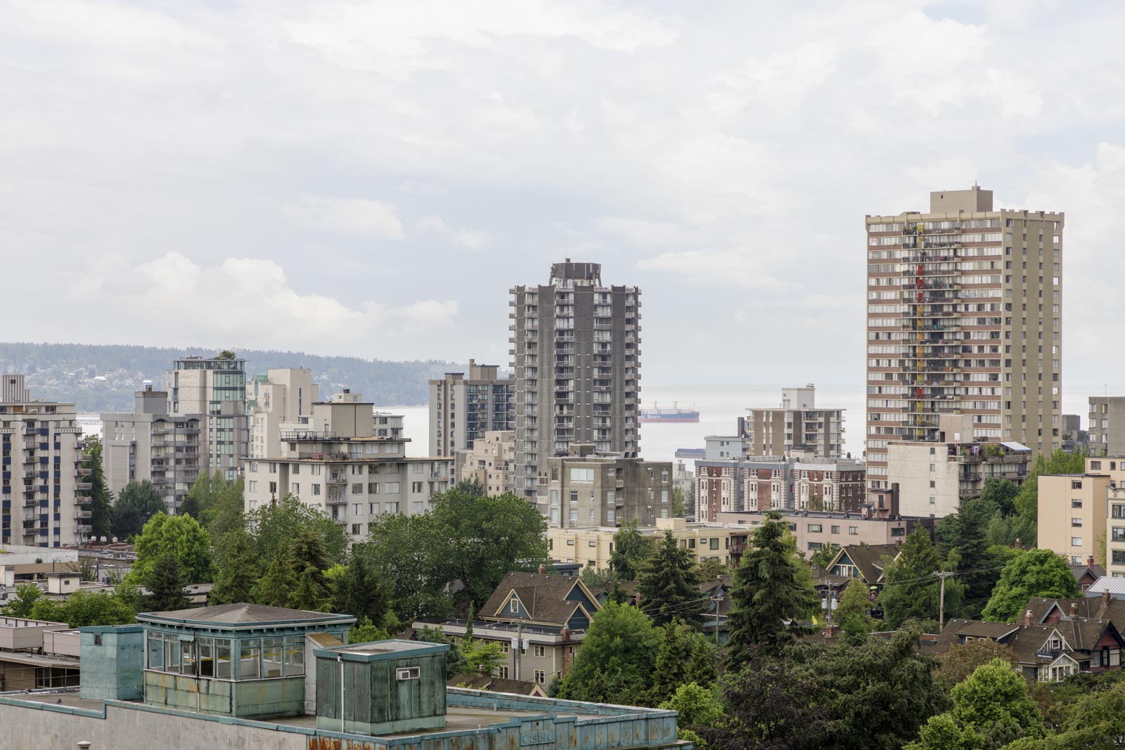 View of a cityscape featuring mid- and high-rise apartment buildings with trees and houses in the foreground under a cloudy sky.