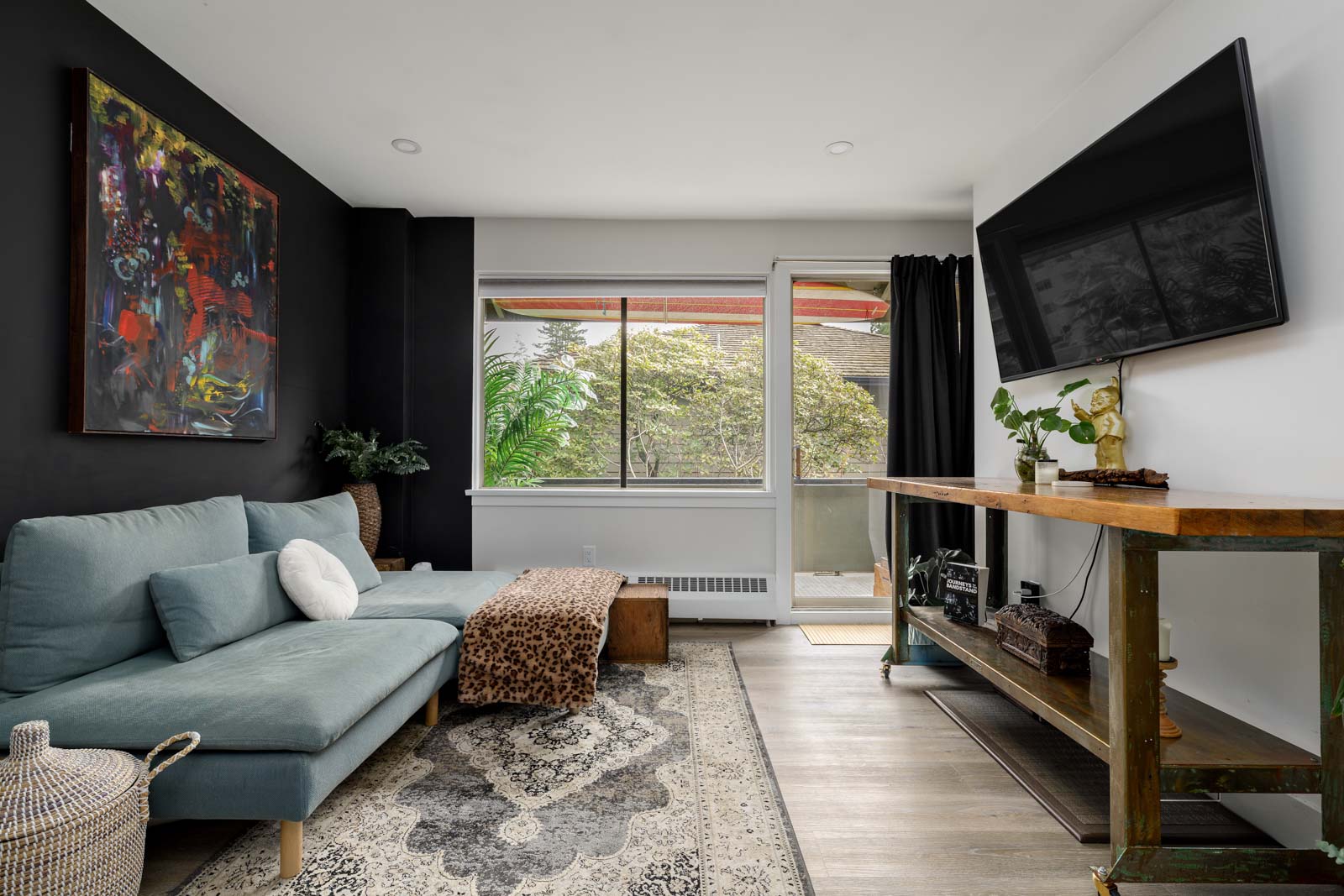 Modern living room with a gray sofa, abstract painting, and a flat-screen TV. Large window shows greenery outside. Decor includes a patterned rug and potted plants.