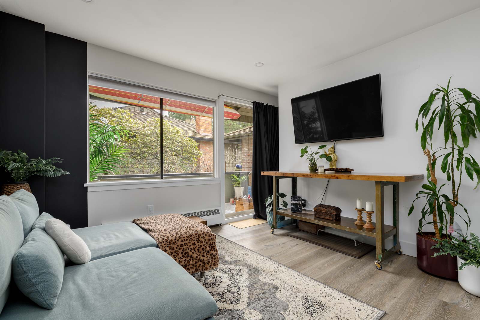 A cozy living room with a sectional sofa, a TV mounted on the wall, a wooden console table, potted plants, a large window, and a glass door leading to a patio.