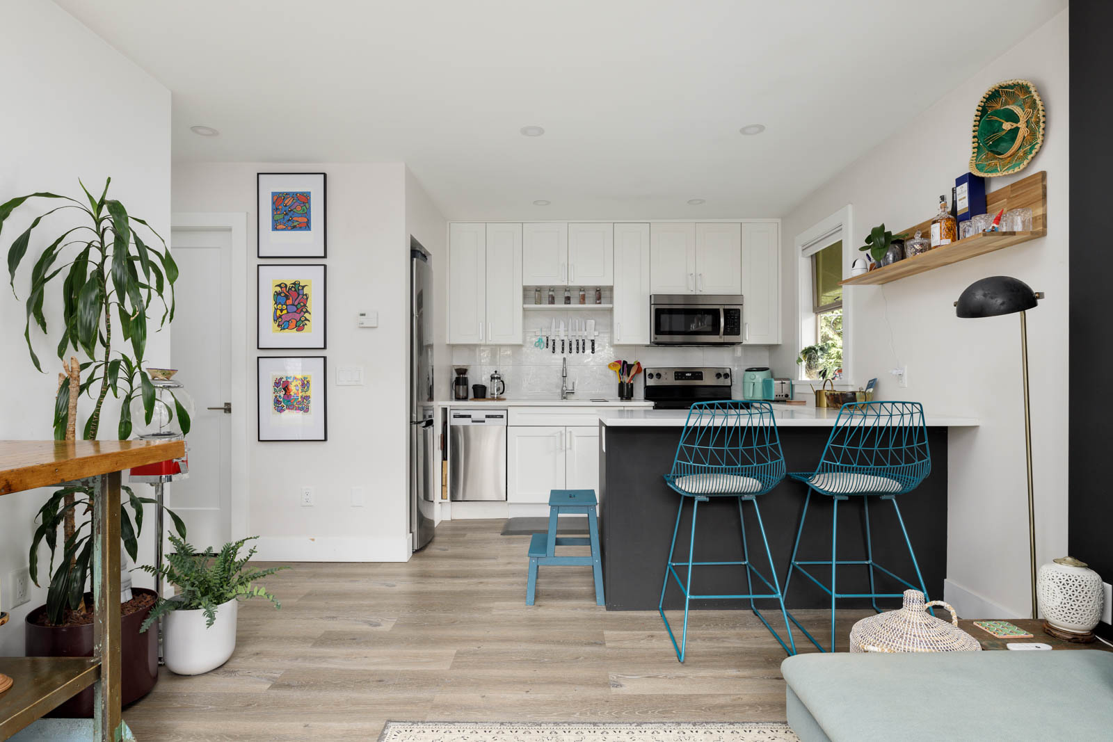 Modern open kitchen with white cabinets, a black island with two teal wire stools, and wooden shelves. Green plants and framed artwork decorate the space. Light wood flooring throughout.