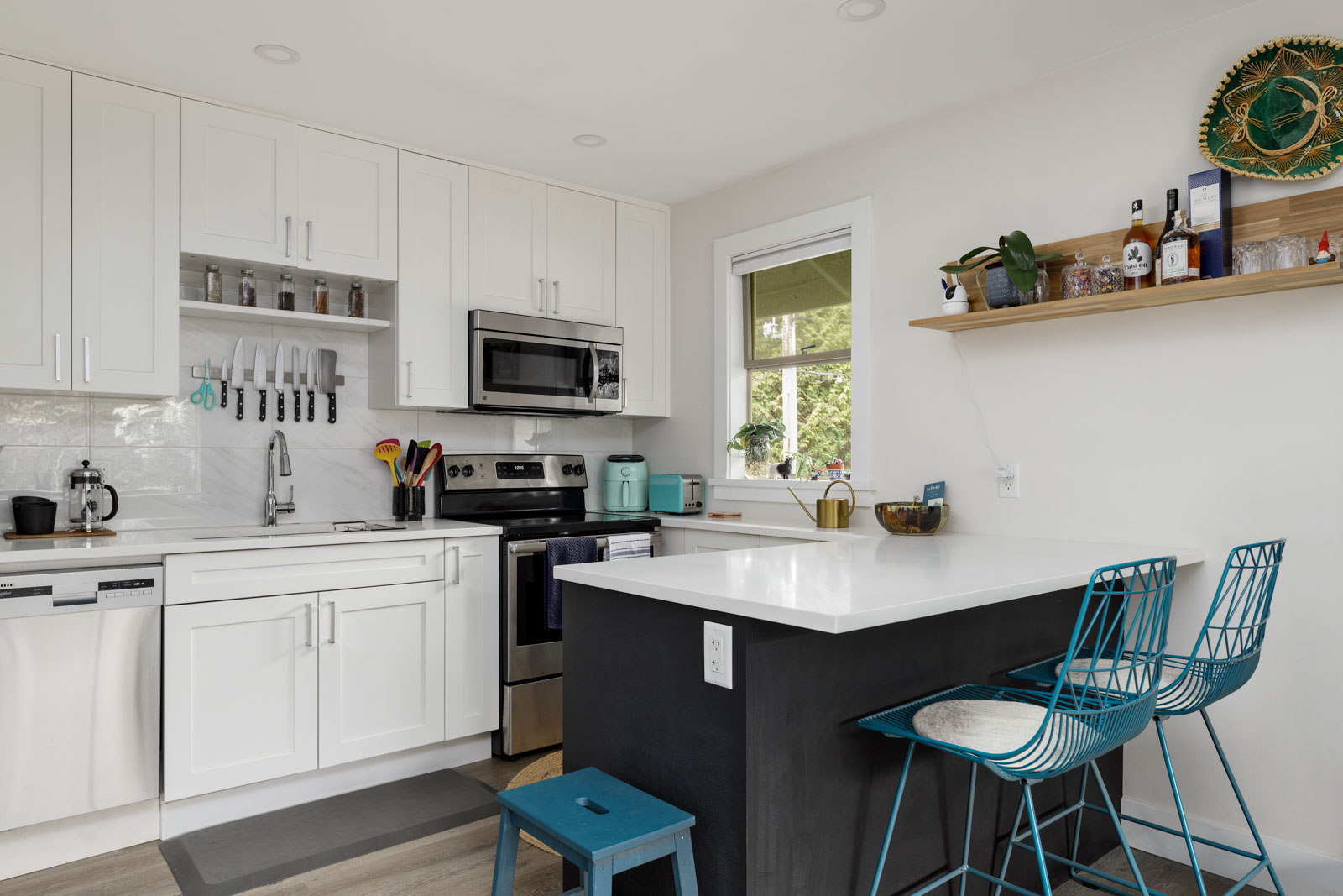 Modern kitchen with white cabinets, stainless steel appliances, and a small island. A shelf holds decor items and a few bottles. Two turquoise stools are at the island. A window brings in natural light.