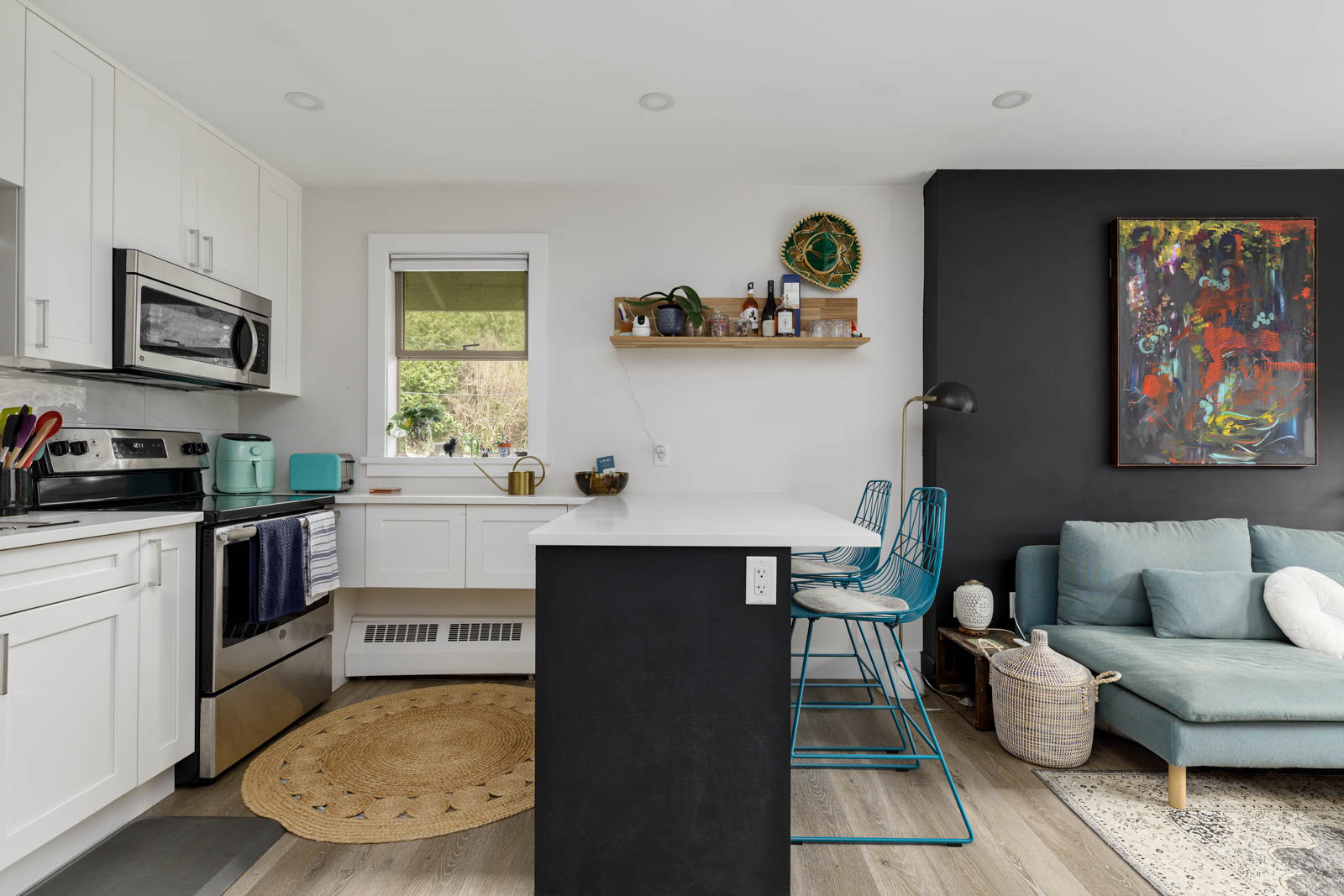 Modern kitchen and living area with white cabinets, black countertops, a teal kitchen appliance, abstract art, and a blue sofa. Two bar stools are at the counter and decor items are on the shelves.