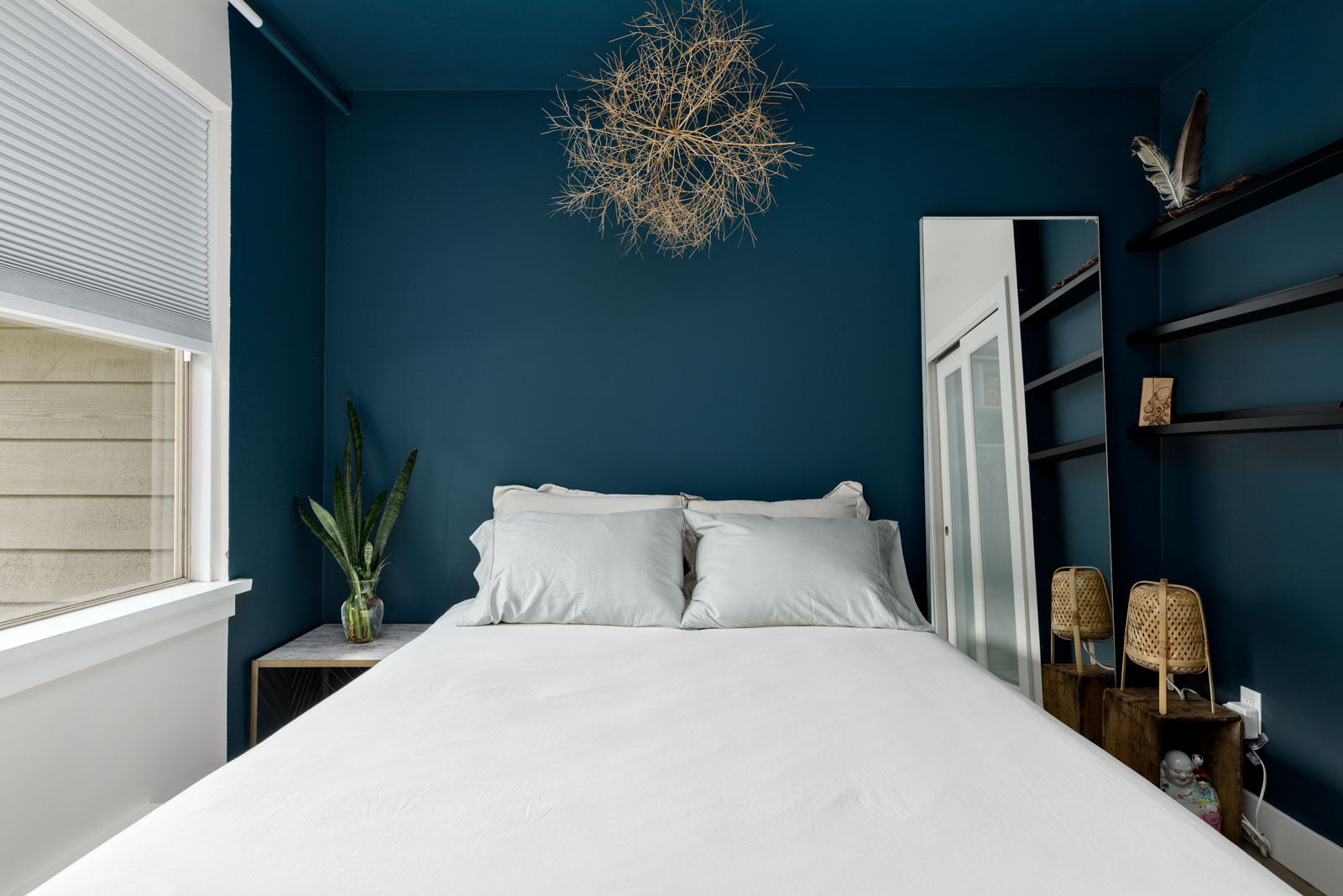 A small bedroom with a blue accent wall, a double bed with white bedding, and decorative hanging branches. A mirror, shelves, and plants adorn the room. Natural light enters through a window.