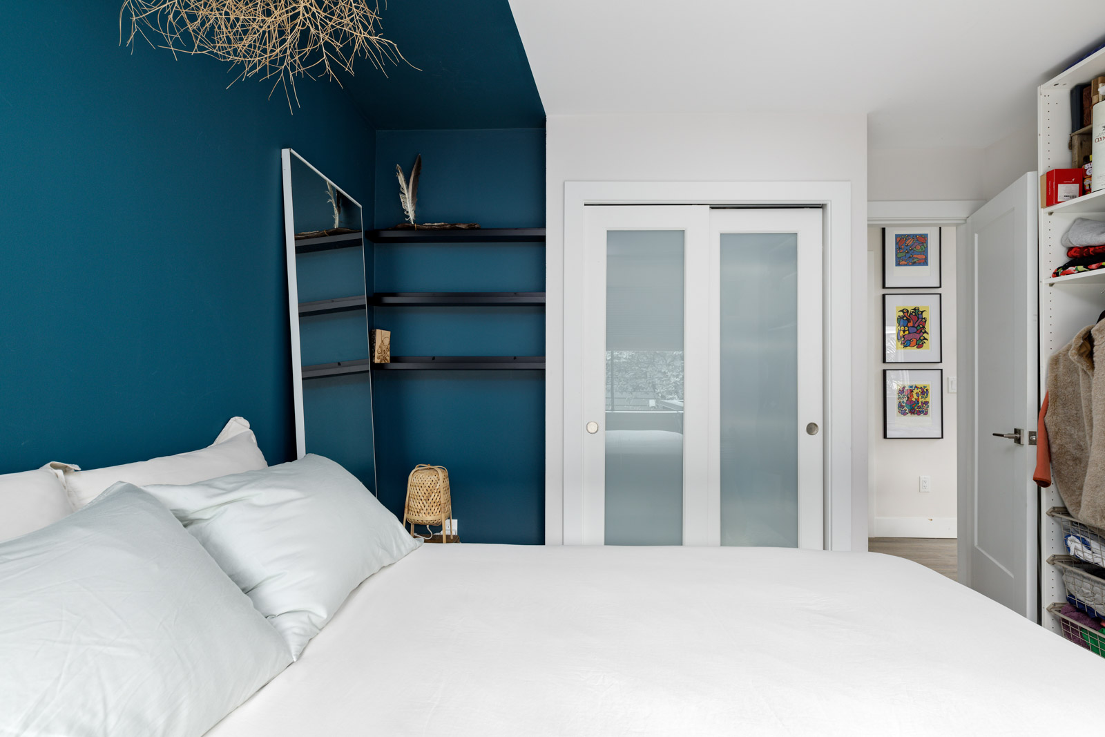 A bedroom with a bed, blue accent wall, white bedding, a floor lamp, a rectangular mirror, a shelving unit, and a sliding closet door.