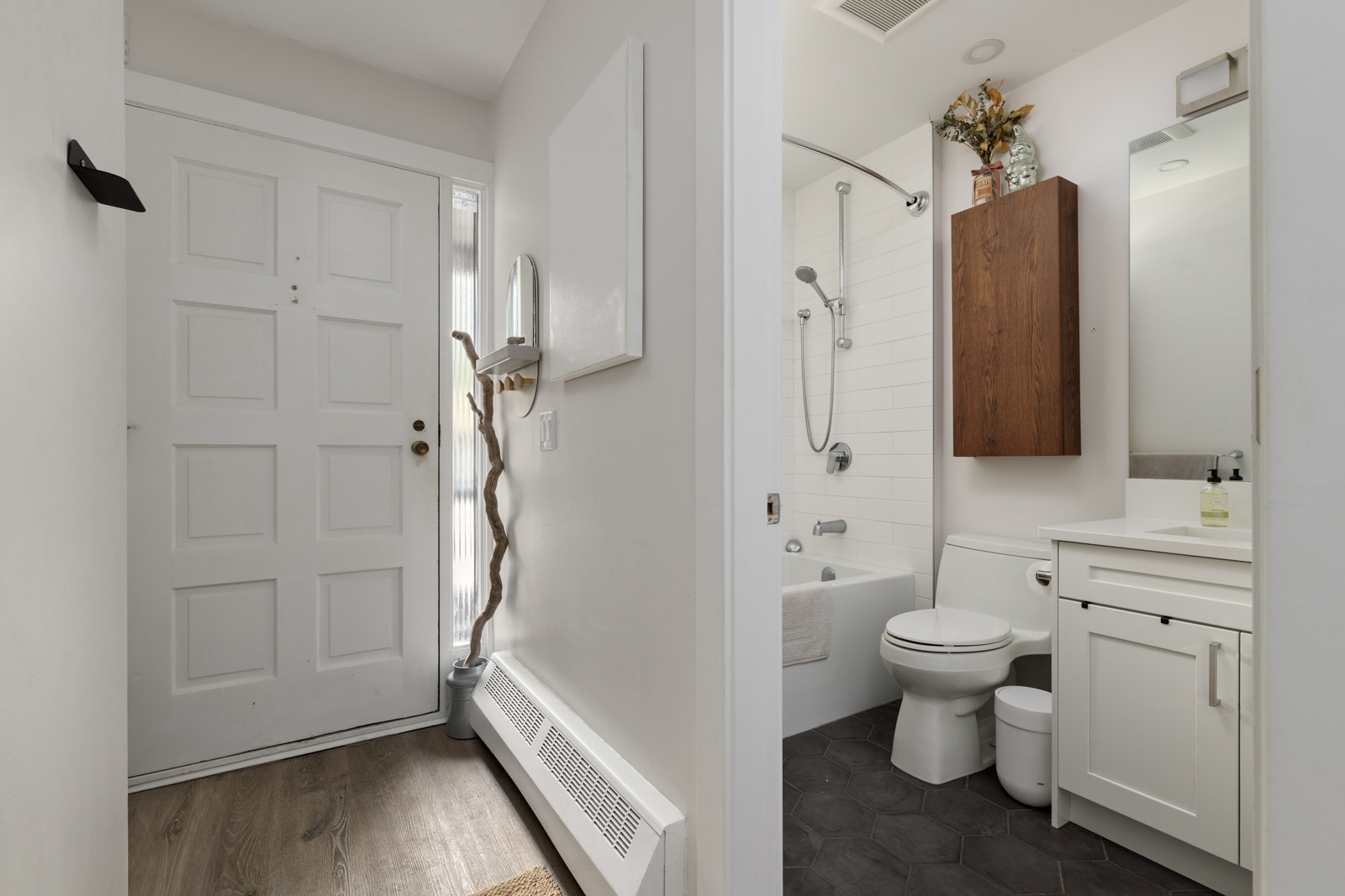 View of a small bathroom with a white door, wooden floor, and a bathroom including a shower, toilet, sink, and wooden cabinet.
