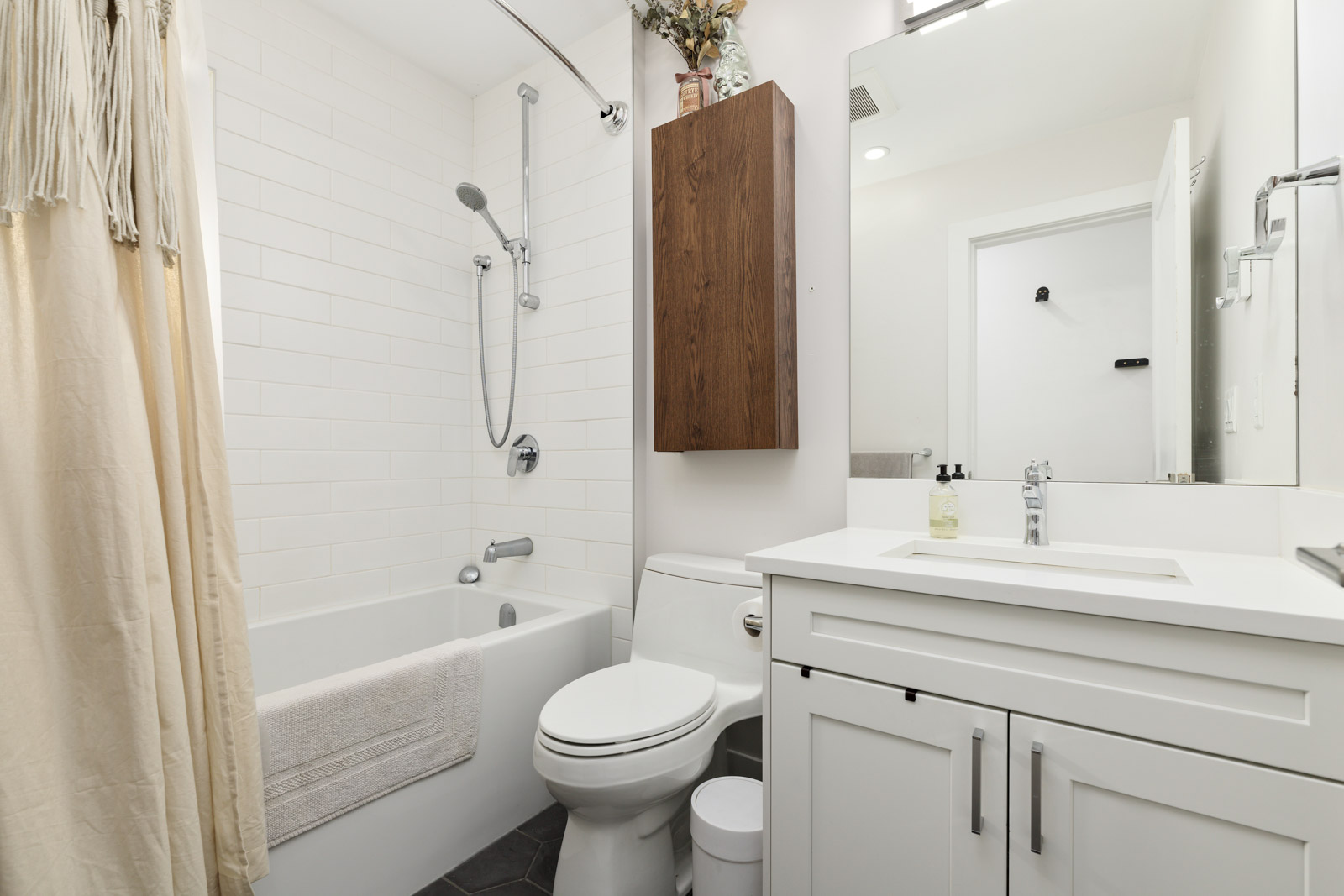 Modern bathroom with white tiles, a bathtub with shower, toilet, and sink. A wooden cabinet and beige shower curtain add warmth. A small plant and soap dispenser are on the counter.