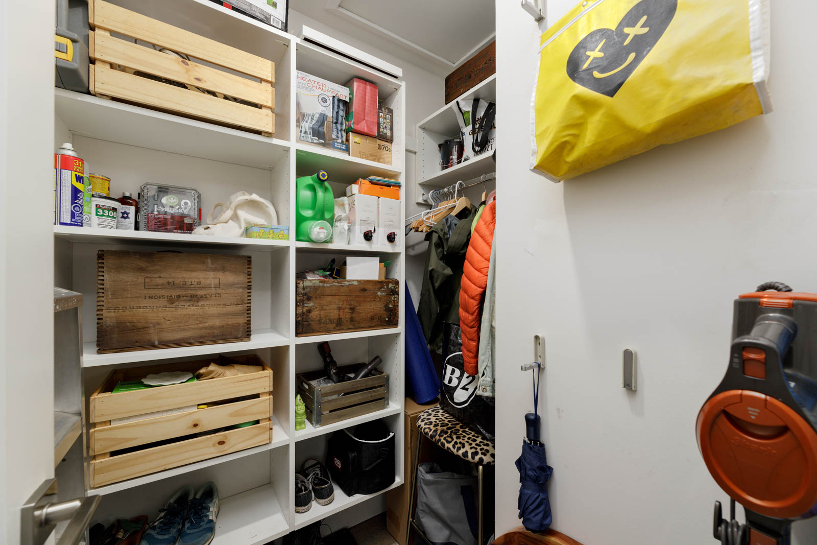 A small closet with shelves holding boxes, cans, and cleaning supplies. Coats and umbrellas hang on a rod; a vacuum is on the floor. A yellow bag with a face design hangs on the door.