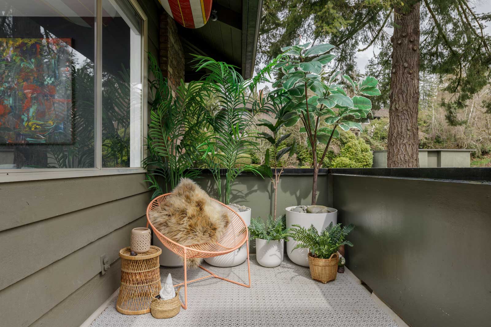 A cozy balcony with a fur-covered chair, green plants in pots, and wicker furniture. A large tree is visible outside.