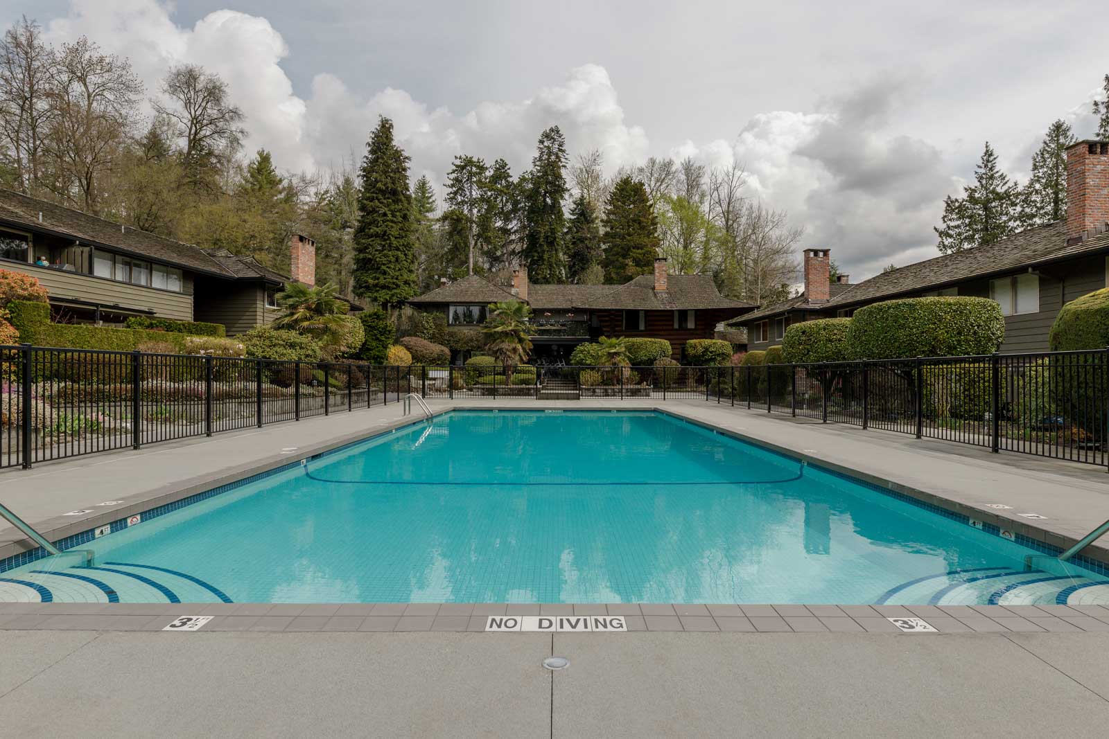Outdoor swimming pool surrounded by black metal fencing, with trees and residential buildings in the background.
