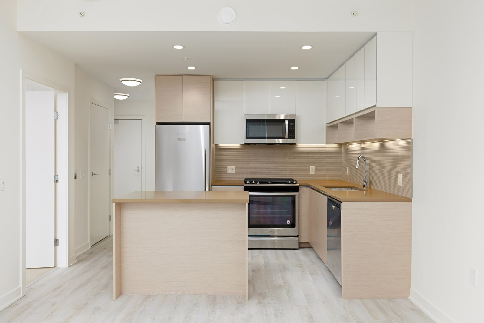 Modern kitchen with light wood cabinets, stainless steel appliances, a built-in oven, microwave, and dishwasher, beige countertops, and light-colored flooring.