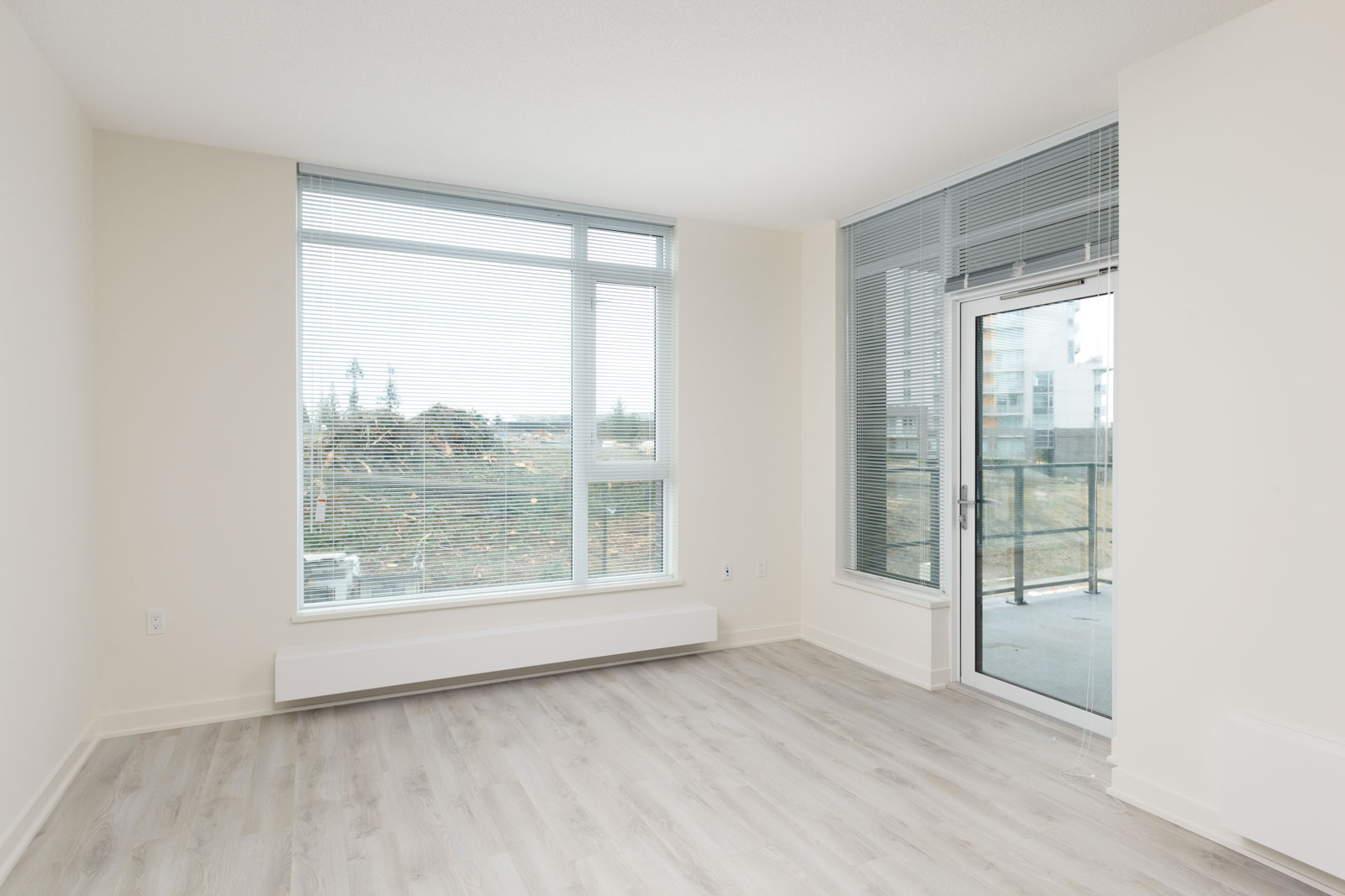 Empty room with light-colored walls and flooring, large window with blinds, and glass door leading to a balcony with view of buildings and trees outside.