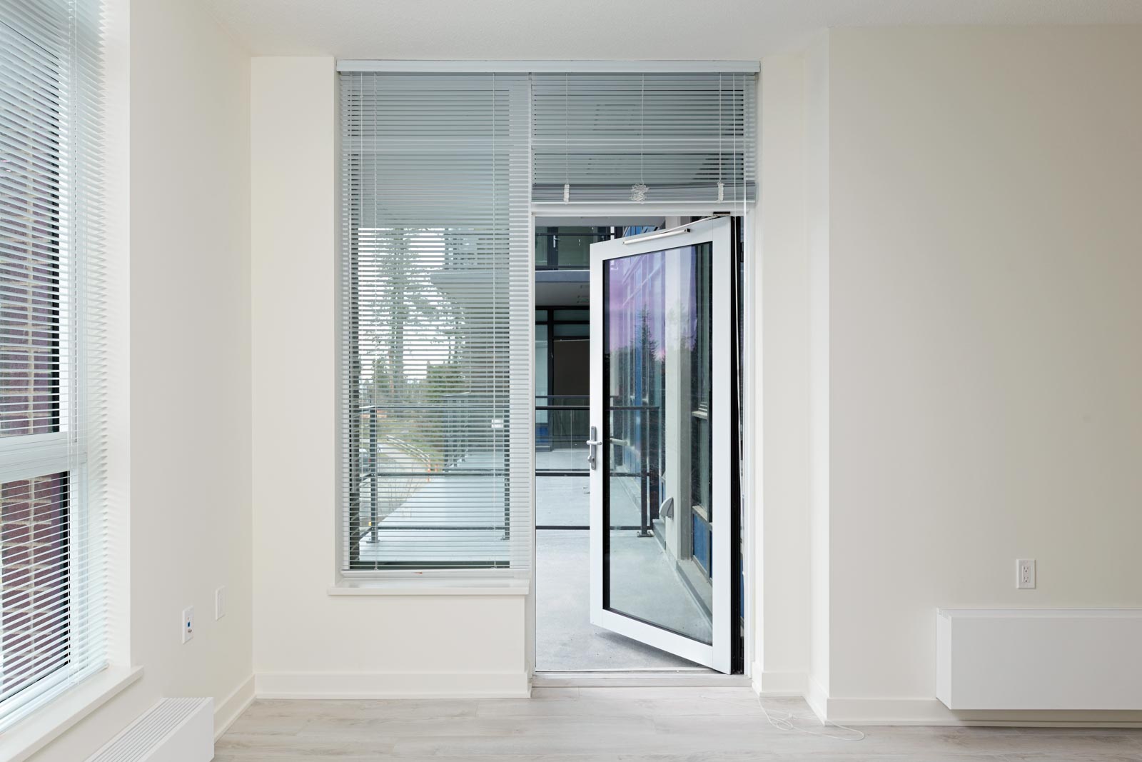 A modern, unfurnished room with large windows and blinds, featuring an open glass door leading to a balcony.