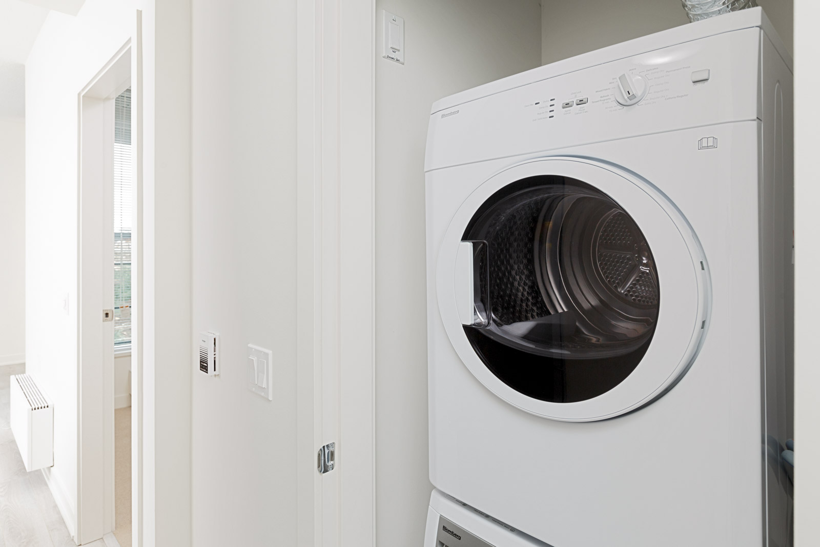 A front-loading white washer and dryer are stacked in a narrow laundry alcove next to a hallway with white walls and a doorway.