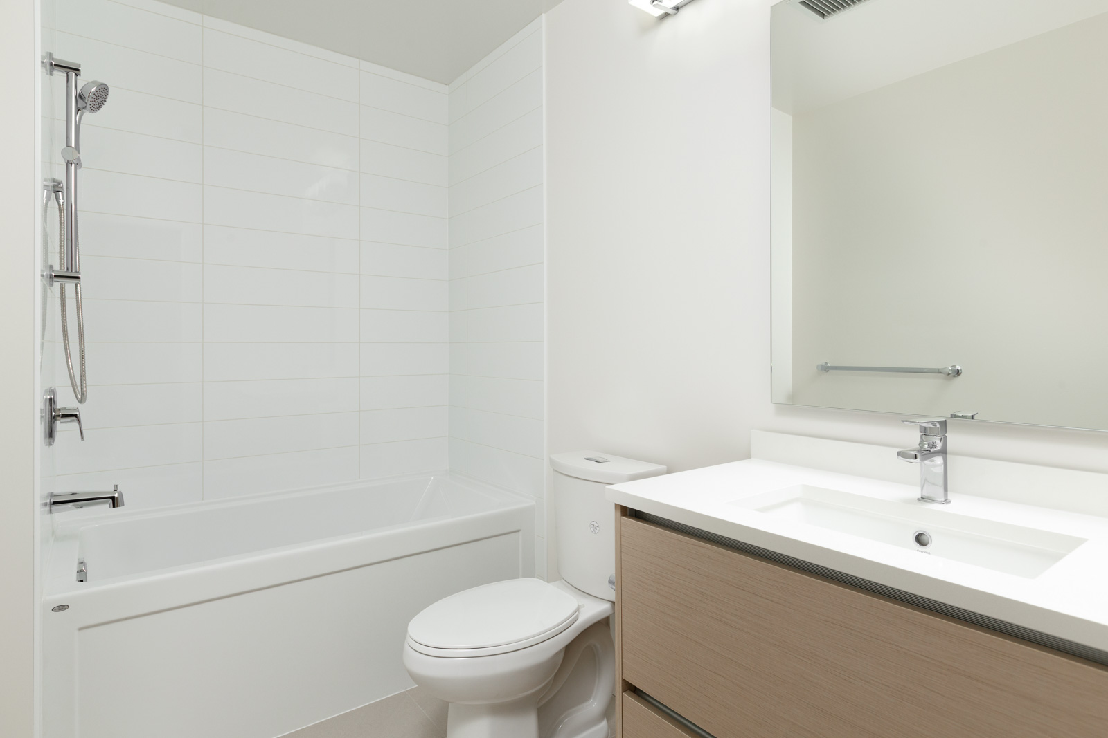 Modern bathroom with a white bathtub and shower, toilet, light wood vanity with sink, large mirror, and white tile walls.