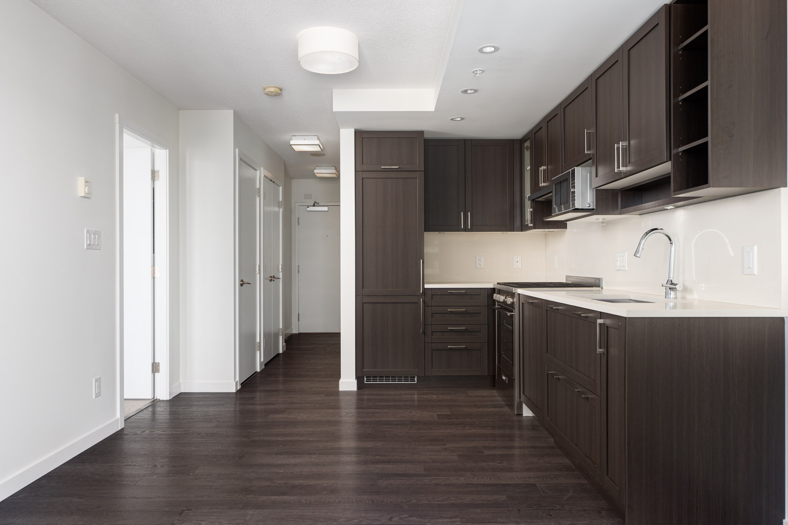 Modern kitchen with dark wood cabinets, stainless steel appliances, white walls, and dark wood flooring, adjacent to a hallway with white doors.