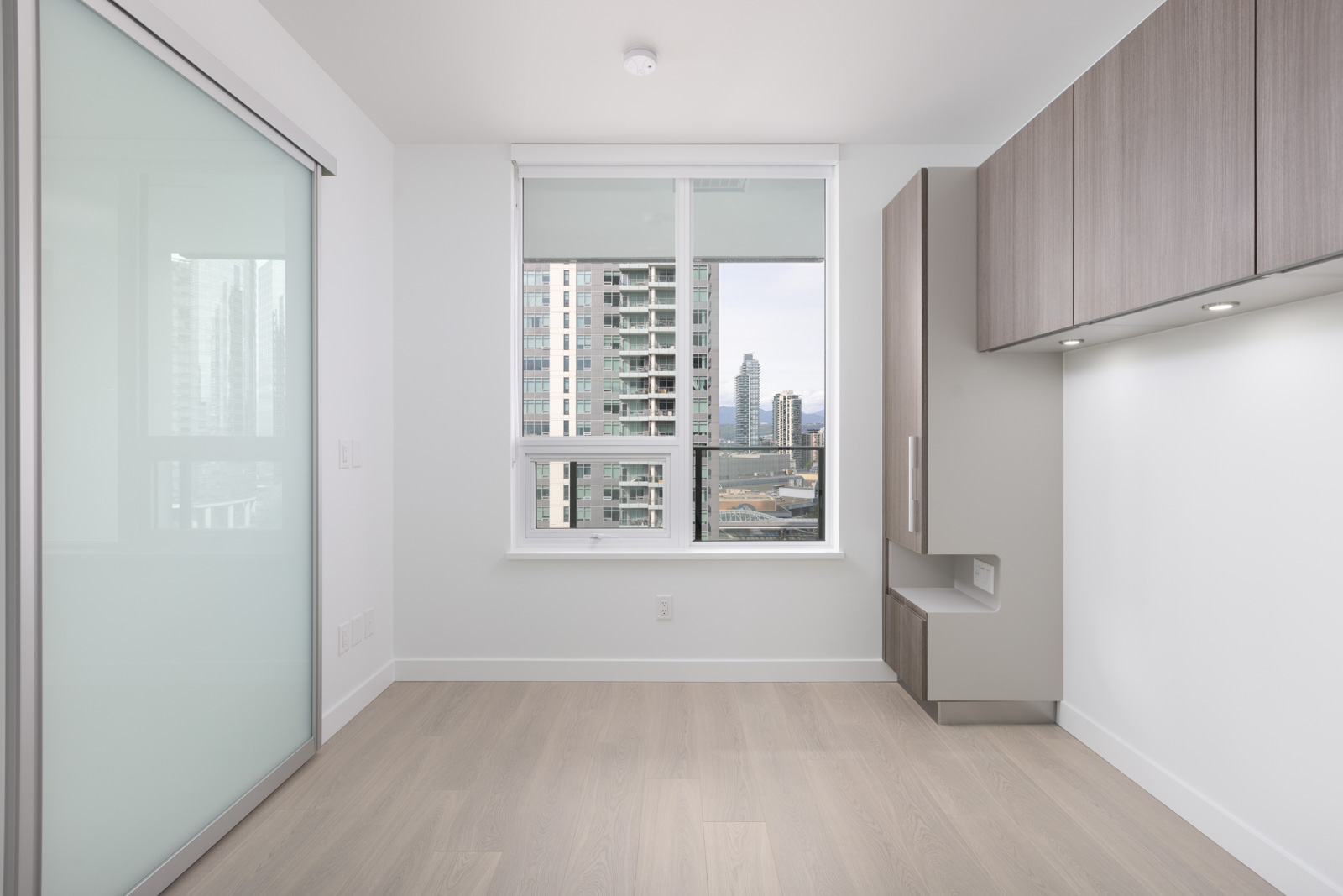 Minimalist empty room with light wood flooring, built-in cabinets, a large window with a city view, and a sliding frosted glass door.