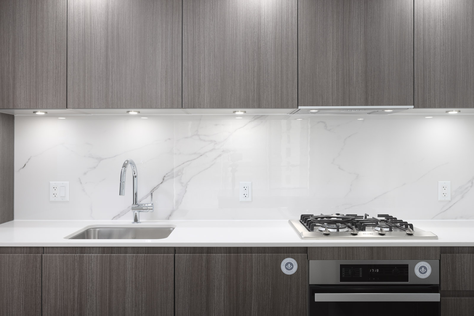 Modern kitchen with gray wood cabinets, marble backsplash, stainless steel sink, and a gas stove under bright built-in lights.