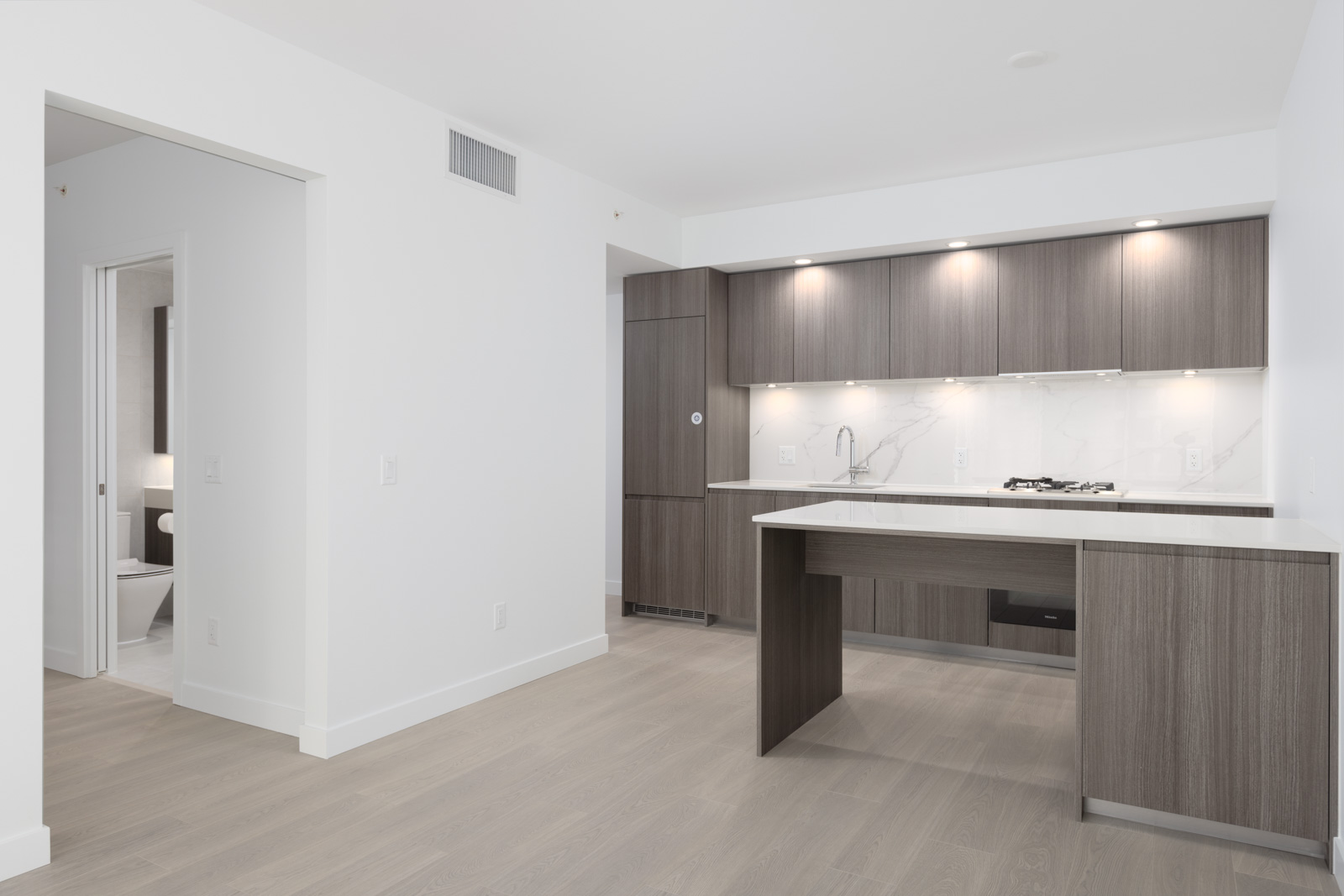 Modern kitchen with wood cabinets, built-in stove, white countertops, and light wood flooring. An open doorway leads to a bathroom with visible toilet.