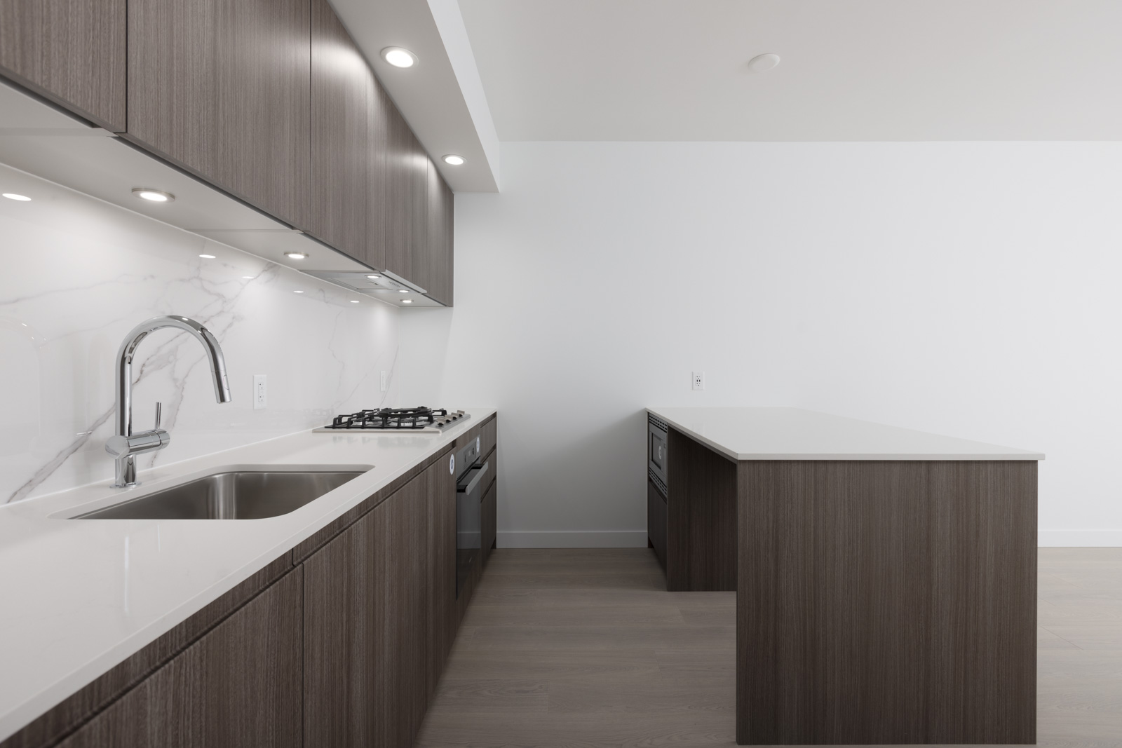 Modern kitchen with dark wood cabinets, white countertops, stainless steel sink, gas stove, under-cabinet lighting, and an island against a white wall.