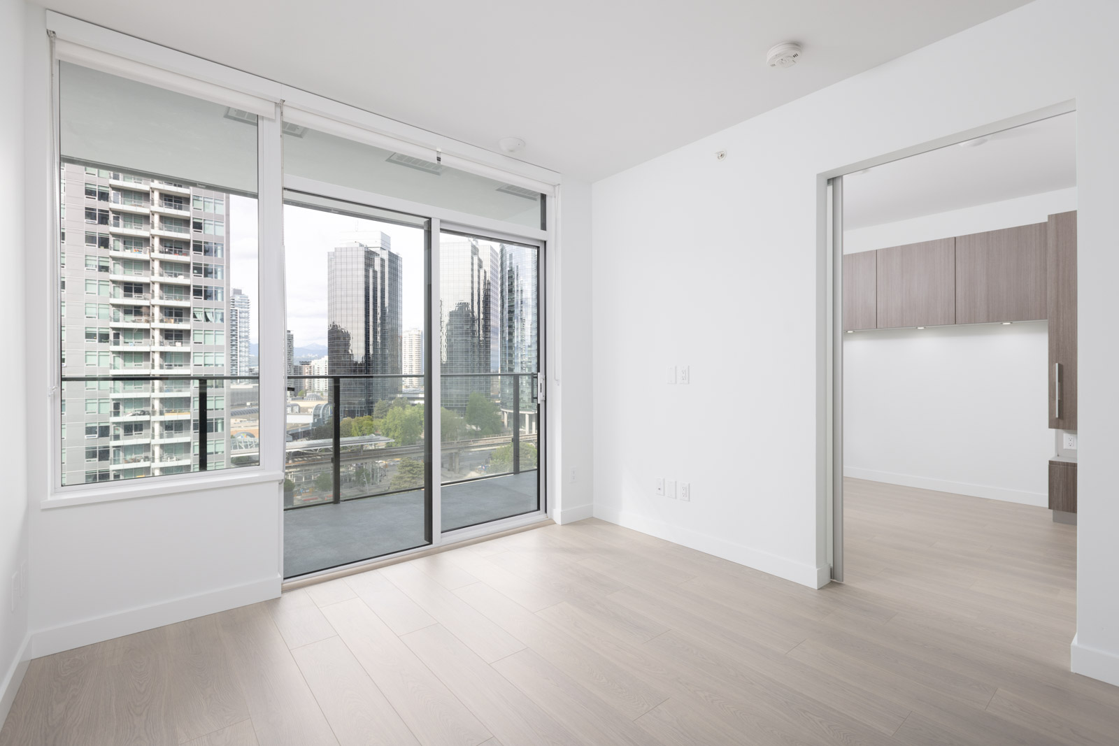 Modern apartment with light wood floors, large windows, sliding glass door to a balcony, and partial view of a kitchen and city buildings outside.