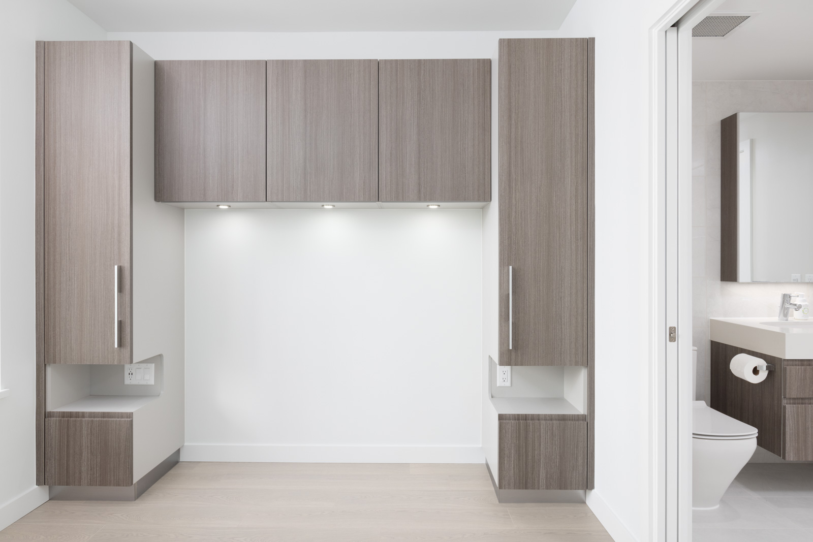 Modern, minimalist laundry or utility area with wood cabinetry, white walls, under-cabinet lighting, and an adjacent bathroom visible through an open door.