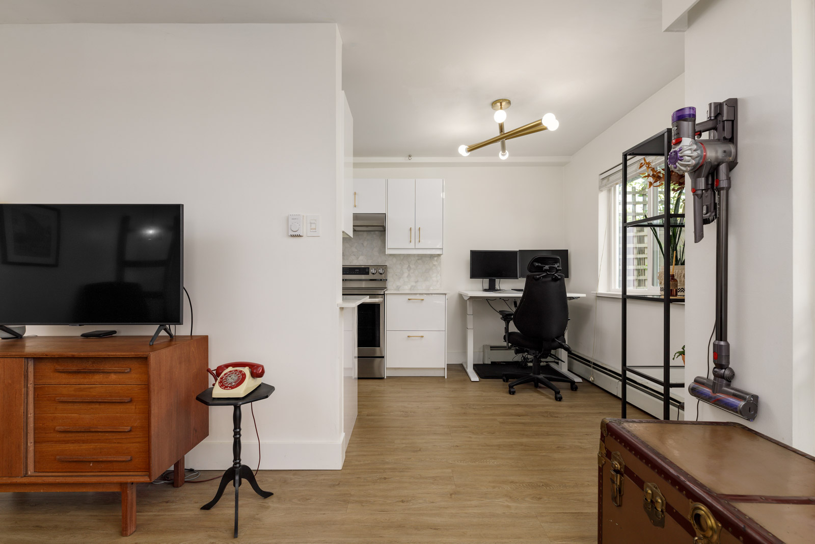A small modern apartment with a TV on a wooden stand, a kitchen area, a computer desk with a chair, a vacuum mounted on the wall, and a trunk in the corner.