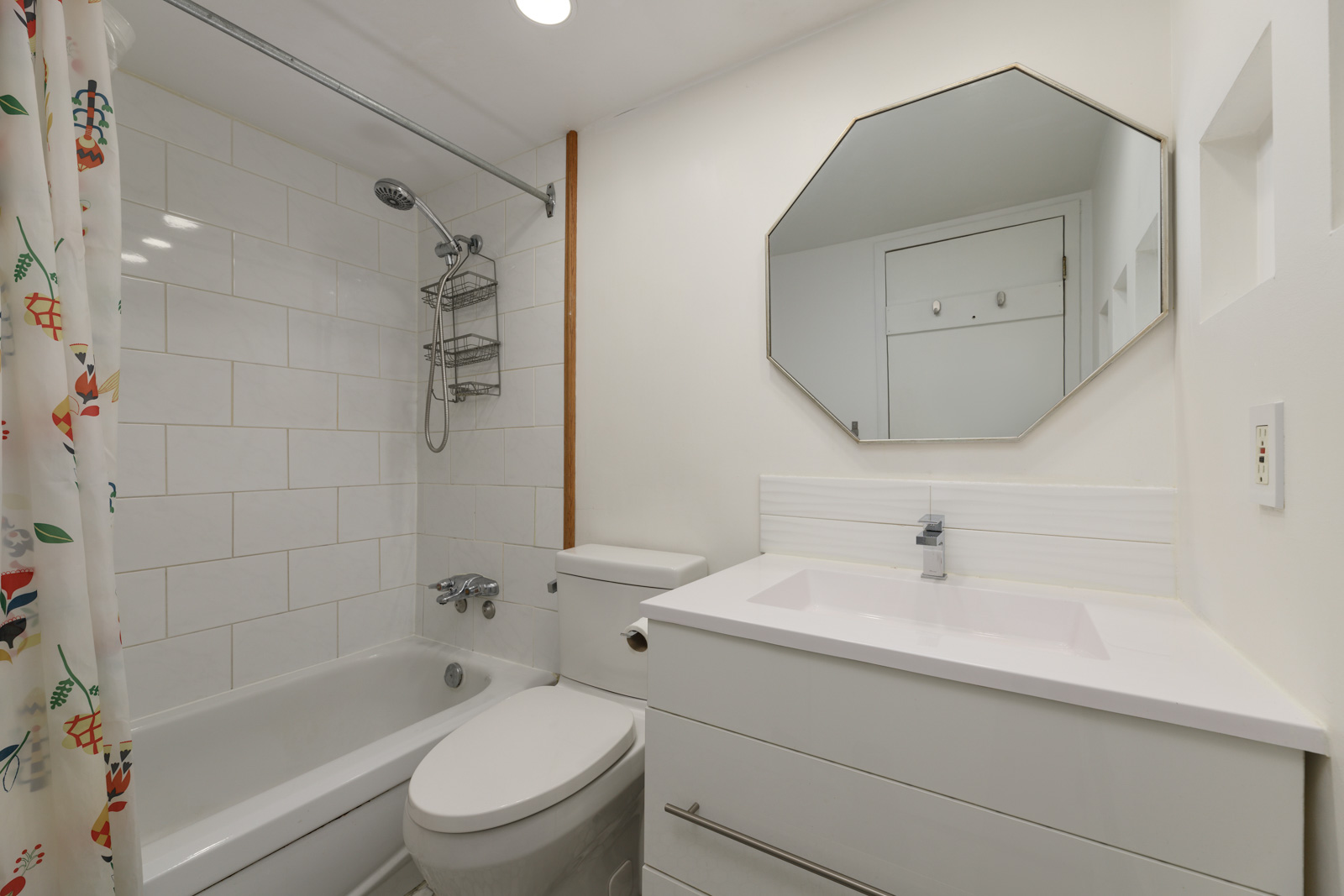 White bathroom with a bathtub-shower combo, colorful shower curtain, toilet, modern sink with cabinet, and octagonal mirror above the vanity.