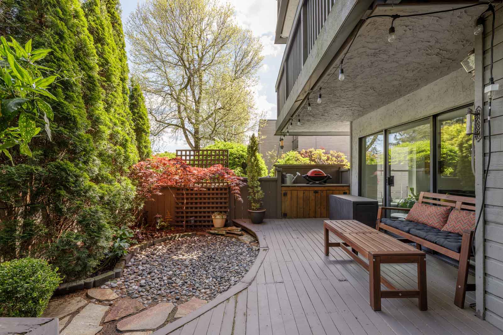 A backyard patio with a wooden deck, outdoor seating, a barbecue grill, potted plants, and a gravel garden area bordered by trees and shrubs.