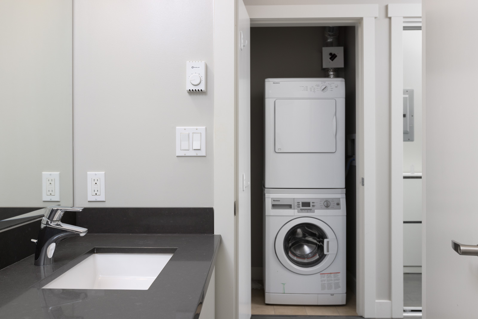 A stacked washer and dryer unit is visible through an open door next to a bathroom with a sink and black countertop.