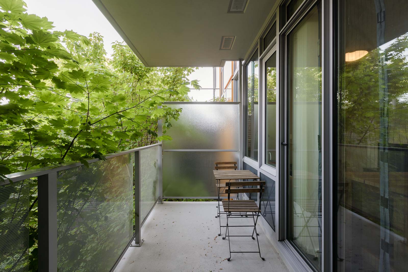 A small apartment balcony with a metal railing, frosted glass privacy screen, and two wooden chairs with a table, surrounded by leafy green trees.