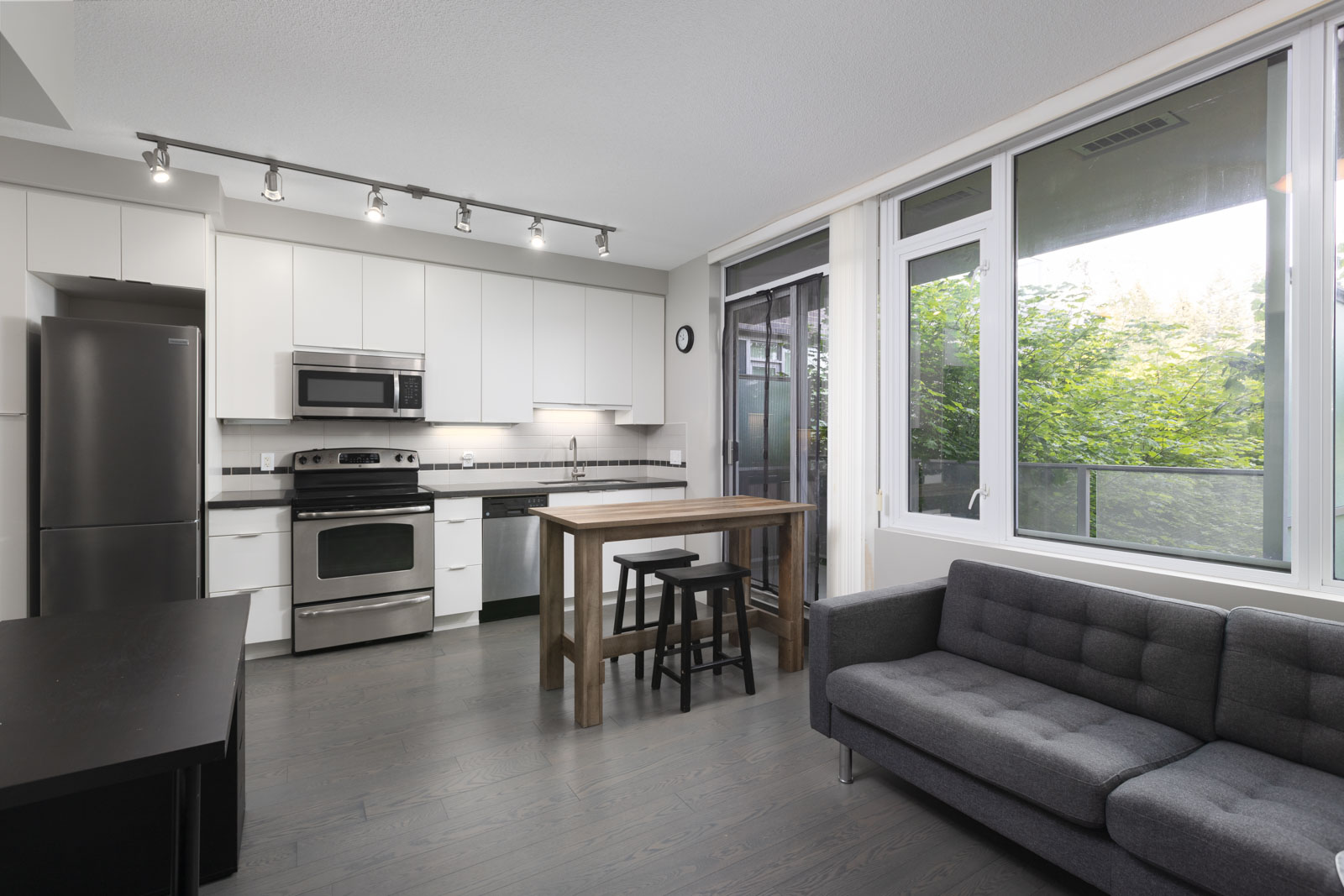 Modern apartment kitchen with stainless steel appliances, white cabinets, a wooden island with stools, gray sofa, large windows, and view of greenery outside.