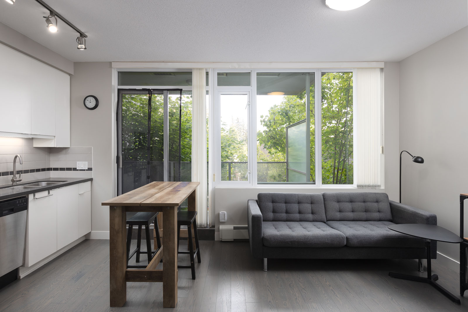 Modern apartment interior with a kitchen, wooden dining table, two stools, gray sofa, floor lamp, and large windows overlooking greenery outside.