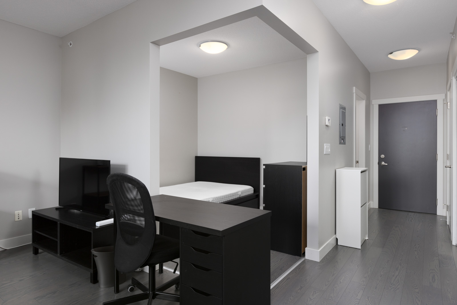 A minimalist studio apartment features a bed in an alcove, a desk with a chair, a TV on a stand, and a dark front door. The decor is modern with neutral tones and clean lines.