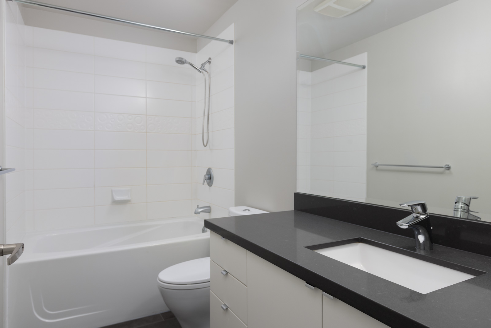Modern bathroom with white bathtub and shower, white toilet, black countertop with sink, large mirror, and white cabinetry.