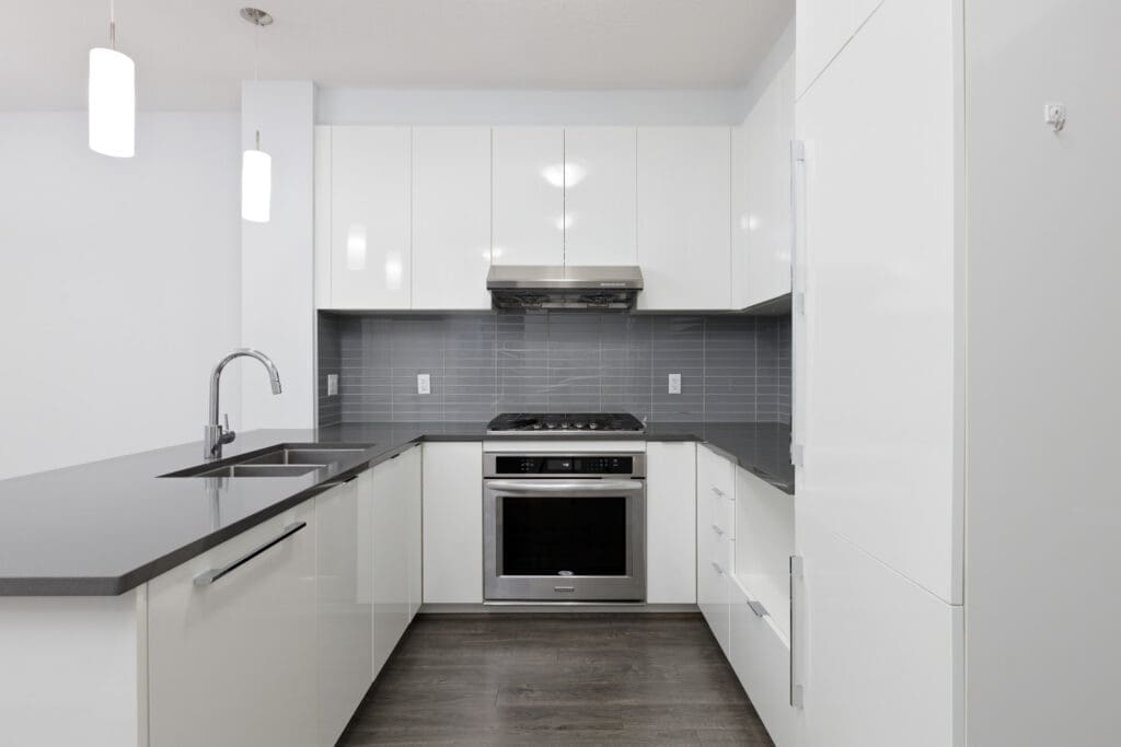 Modern kitchen with white cabinets, gray countertops, stainless steel oven, and gray tile backsplash; pendant lights hang above the sink area.