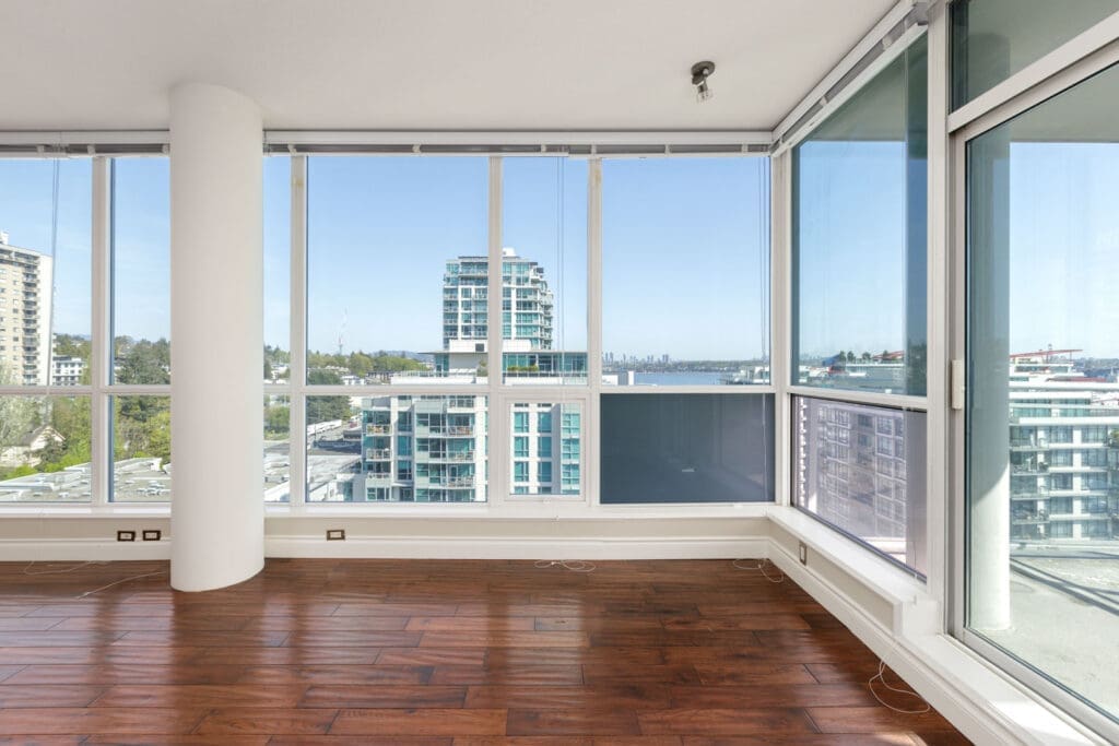 Empty corner room with large floor-to-ceiling windows, hardwood flooring, and a view of nearby buildings and distant water under a clear sky.