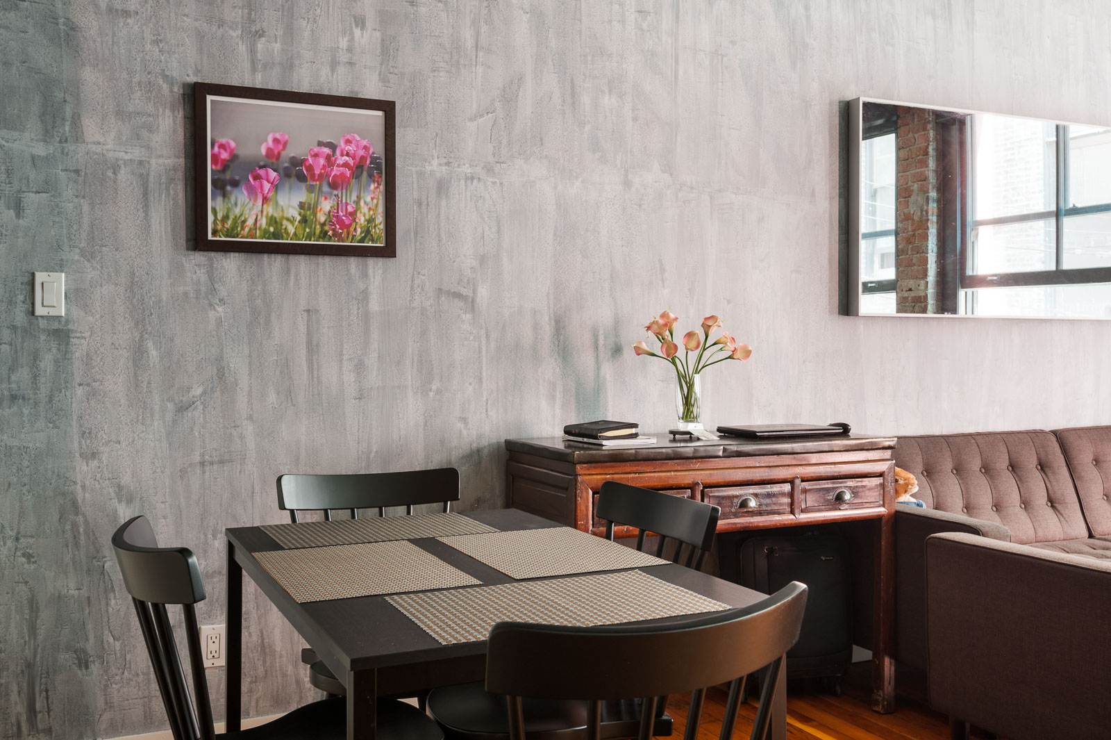 Dining area with a dark table and four chairs, placemats, a sideboard with a vase of flowers, a wall with gray paint, and a framed photo of pink flowers above.