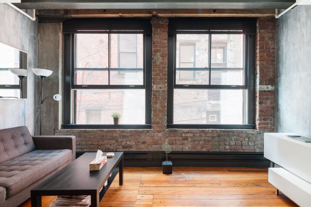 A modern living room with exposed brick walls, large windows, a gray sofa, a dark coffee table, a floor lamp, and wooden floors.