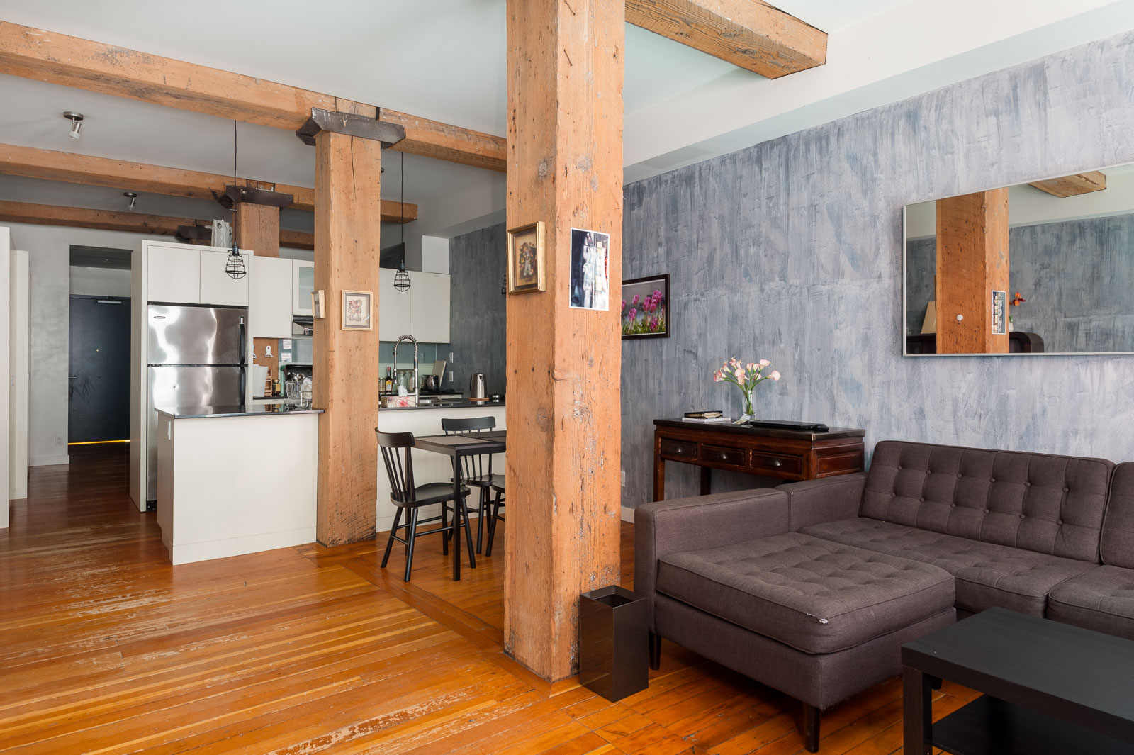Modern loft apartment with exposed wooden beams, open kitchen, gray sectional sofa, small dining area, and decorative wall art.