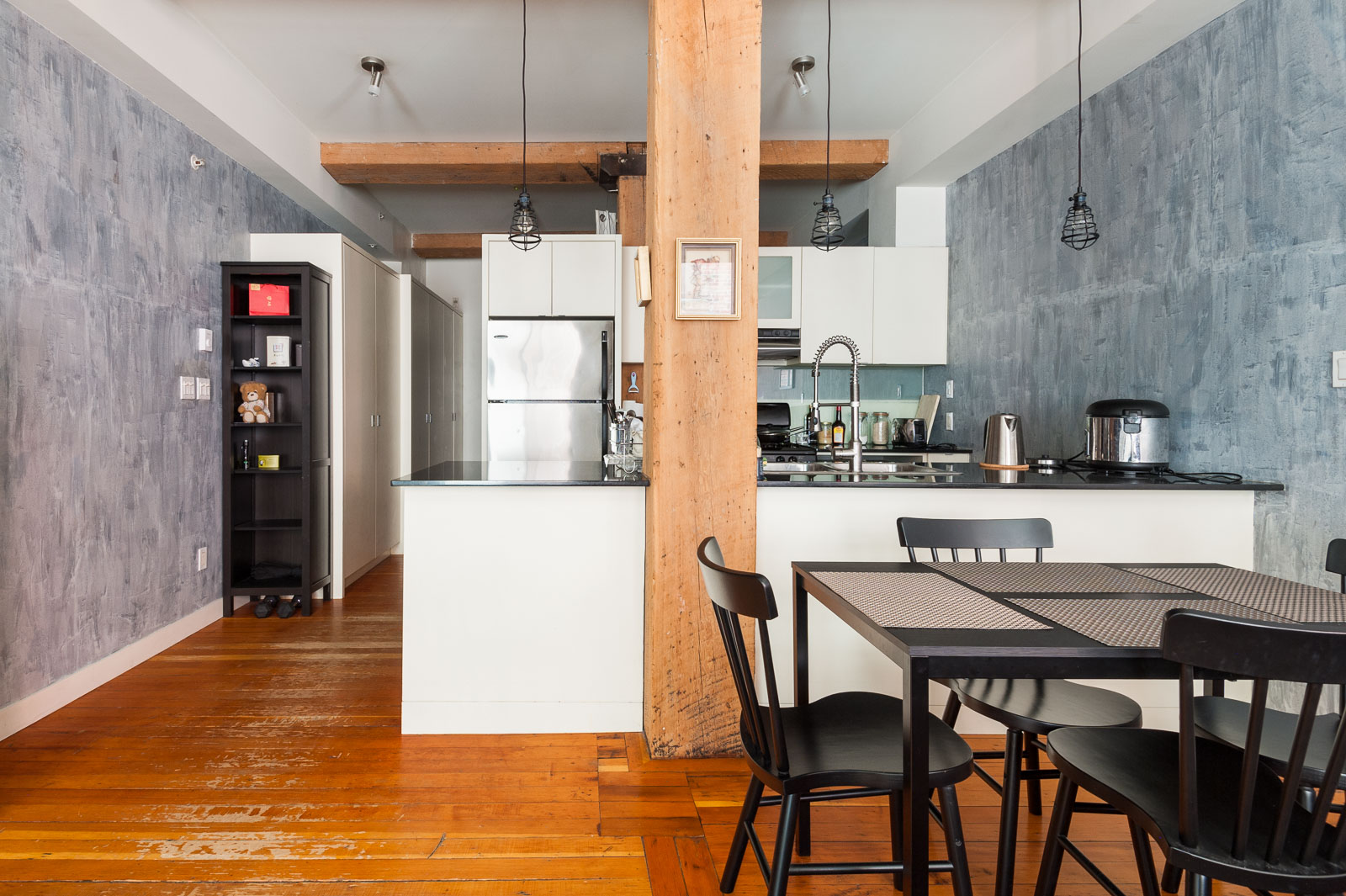 Modern kitchen and dining area with wooden floors, a central wooden beam, white cabinets, stainless steel appliances, and a black dining table with four chairs.