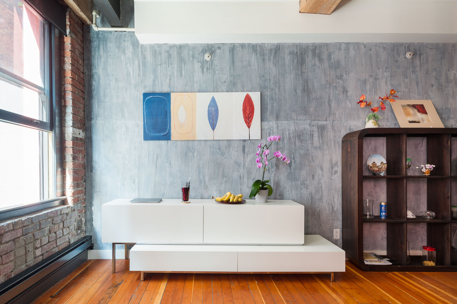 Modern living room with a white sideboard, orchid plant, bowl of bananas, abstract wall art, bookshelf, and exposed brick and wood elements.