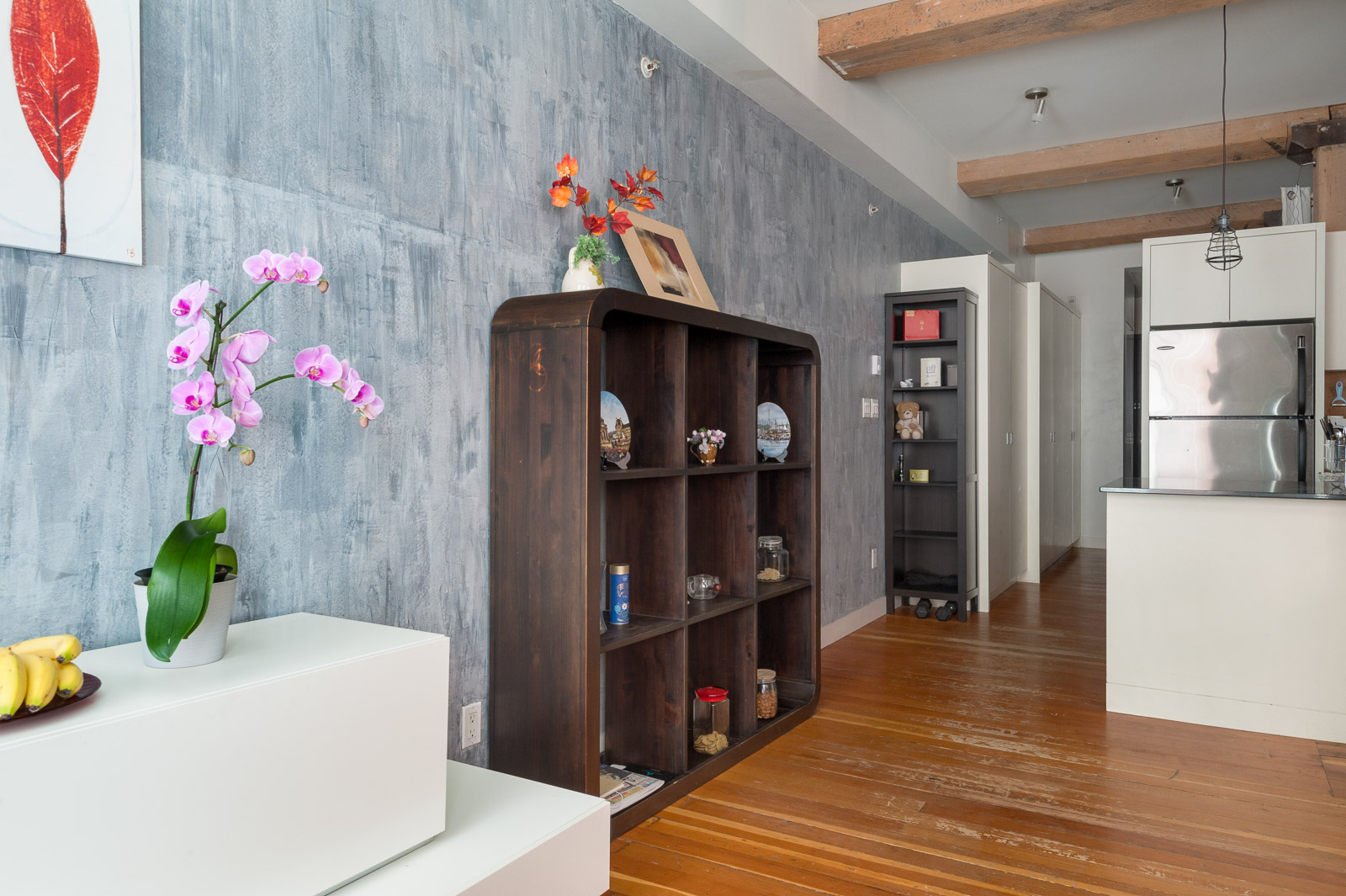 Modern apartment interior with wood flooring, exposed beams, a gray textured wall, open shelves, orchid plant, and a white kitchen with stainless steel refrigerator.