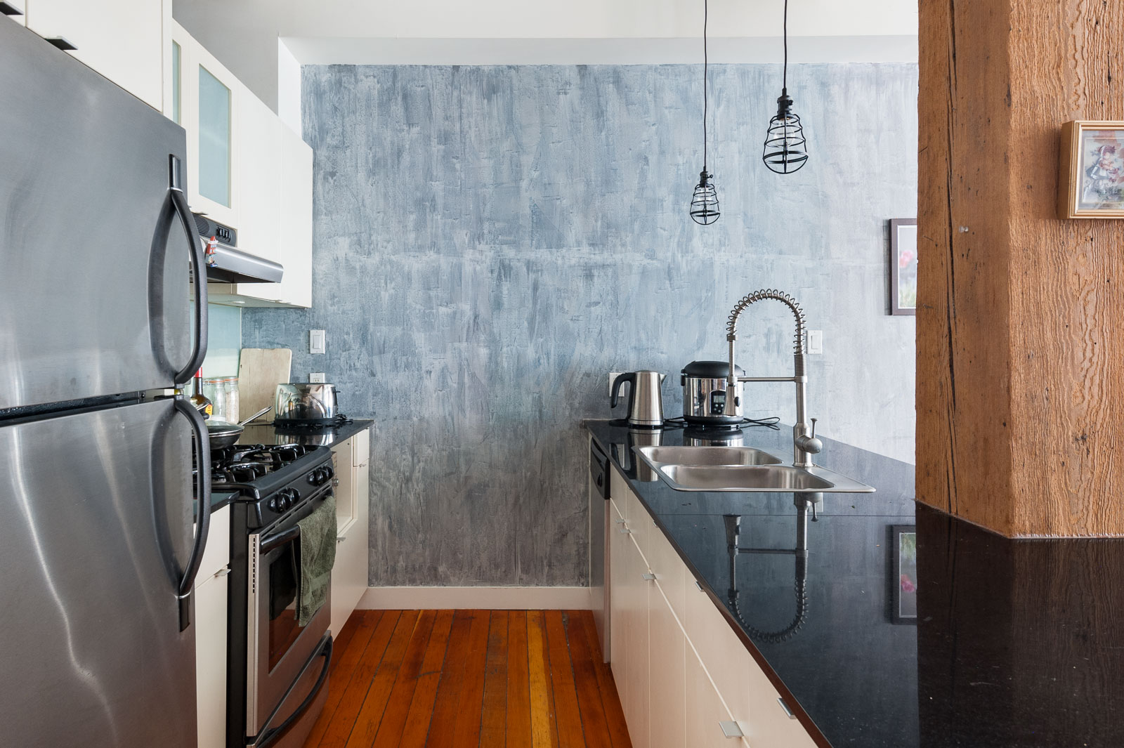 Modern kitchen with stainless steel appliances, a black countertop, a deep sink with a tall faucet, wooden floors, and two pendant lights hanging over the counter.