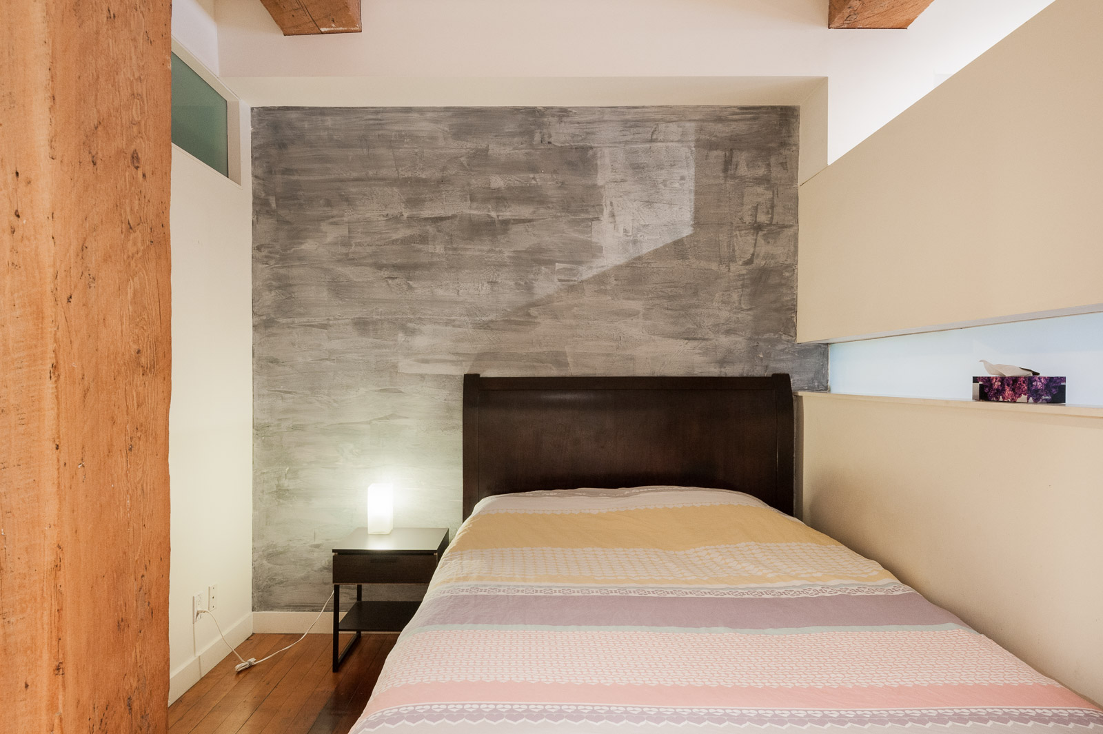 A neatly made single bed with a wooden headboard stands next to a small nightstand and lamp, against a gray textured accent wall in a modern bedroom.